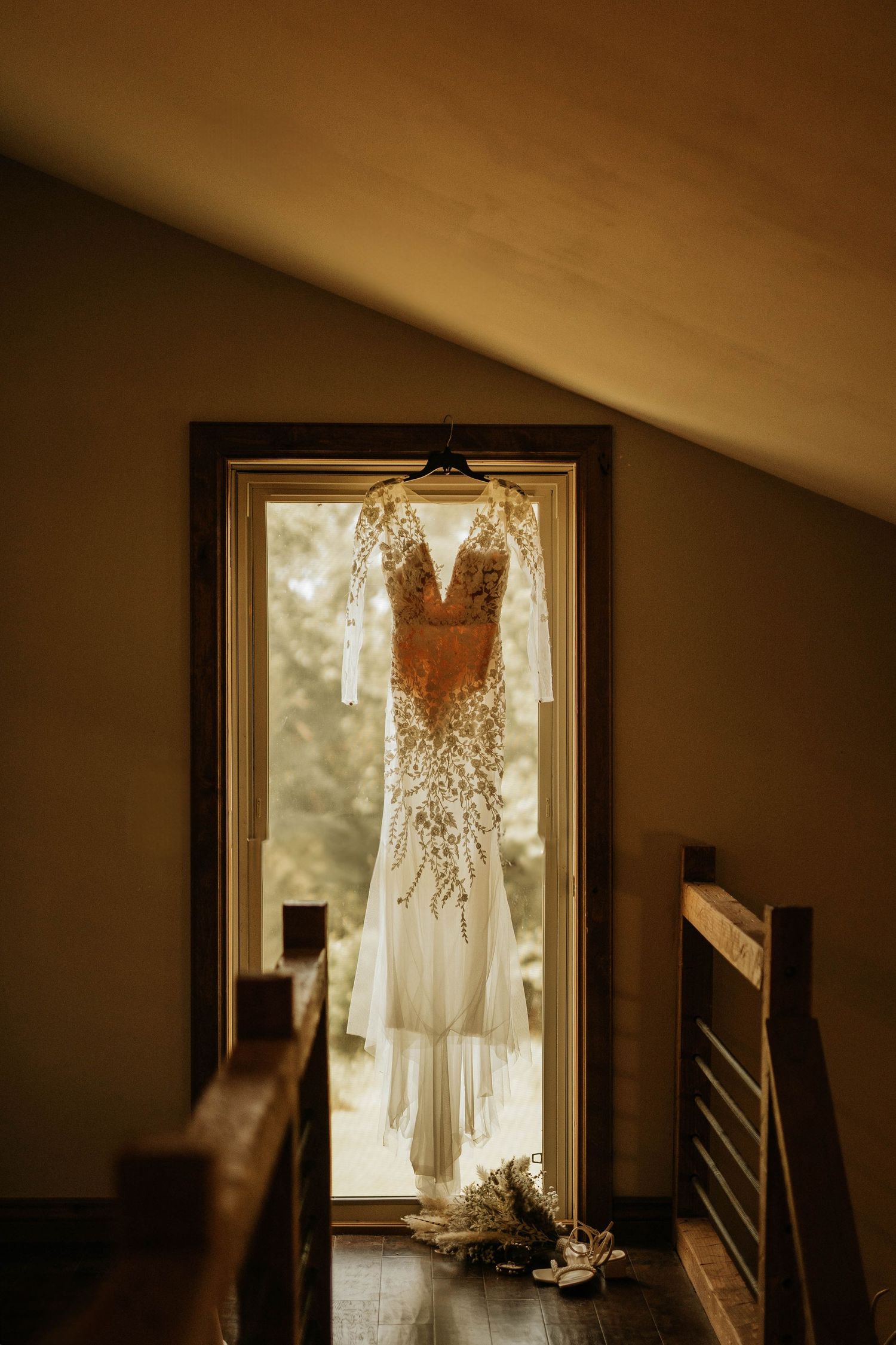 A delicate wedding dress with lace details hangs in a doorway bathed in golden light.