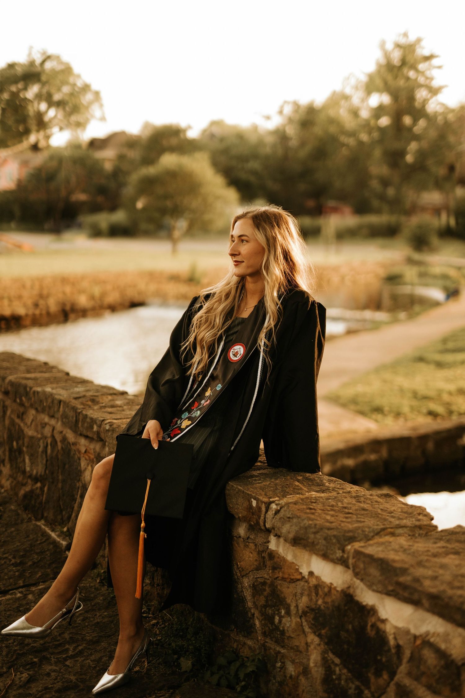 Someone sits on a stone wall at sunset wearing a black coat near a winding path.