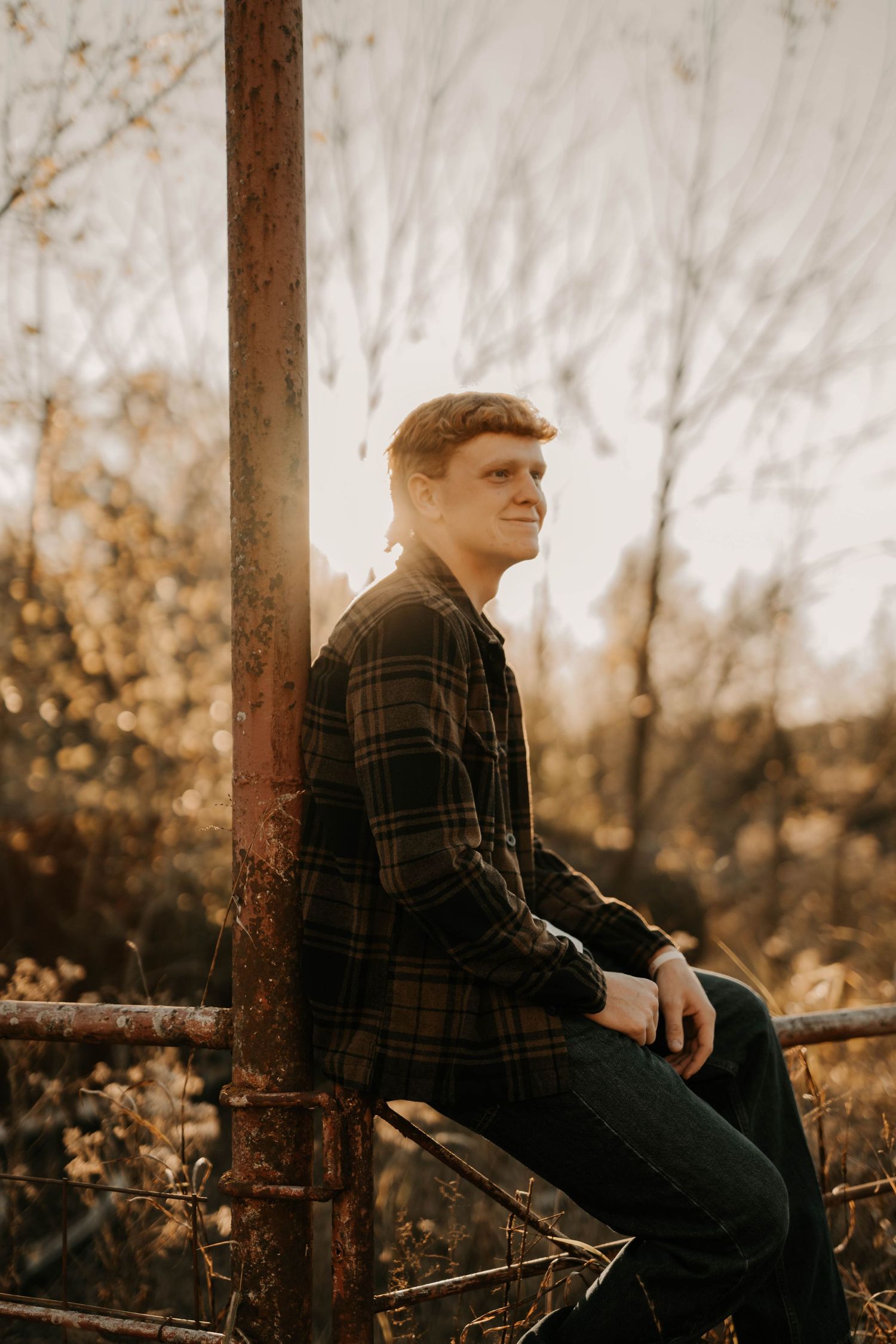 Person in plaid shirt sits on rustic metal fence during golden hour with autumn trees behind.