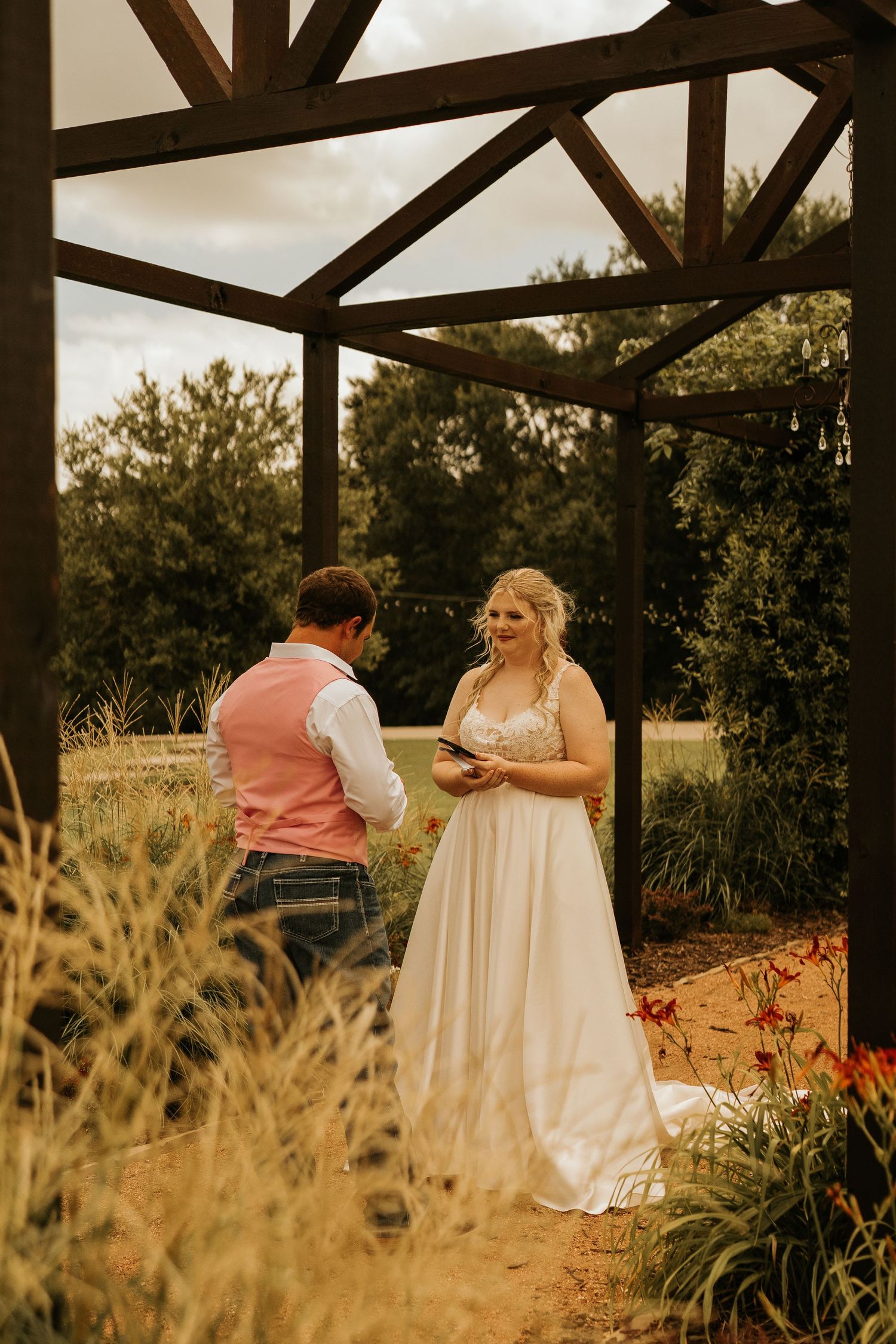 Bride in white gown reads vows to groom in pink vest under rustic wooden pergola surrounded by lush greenery.