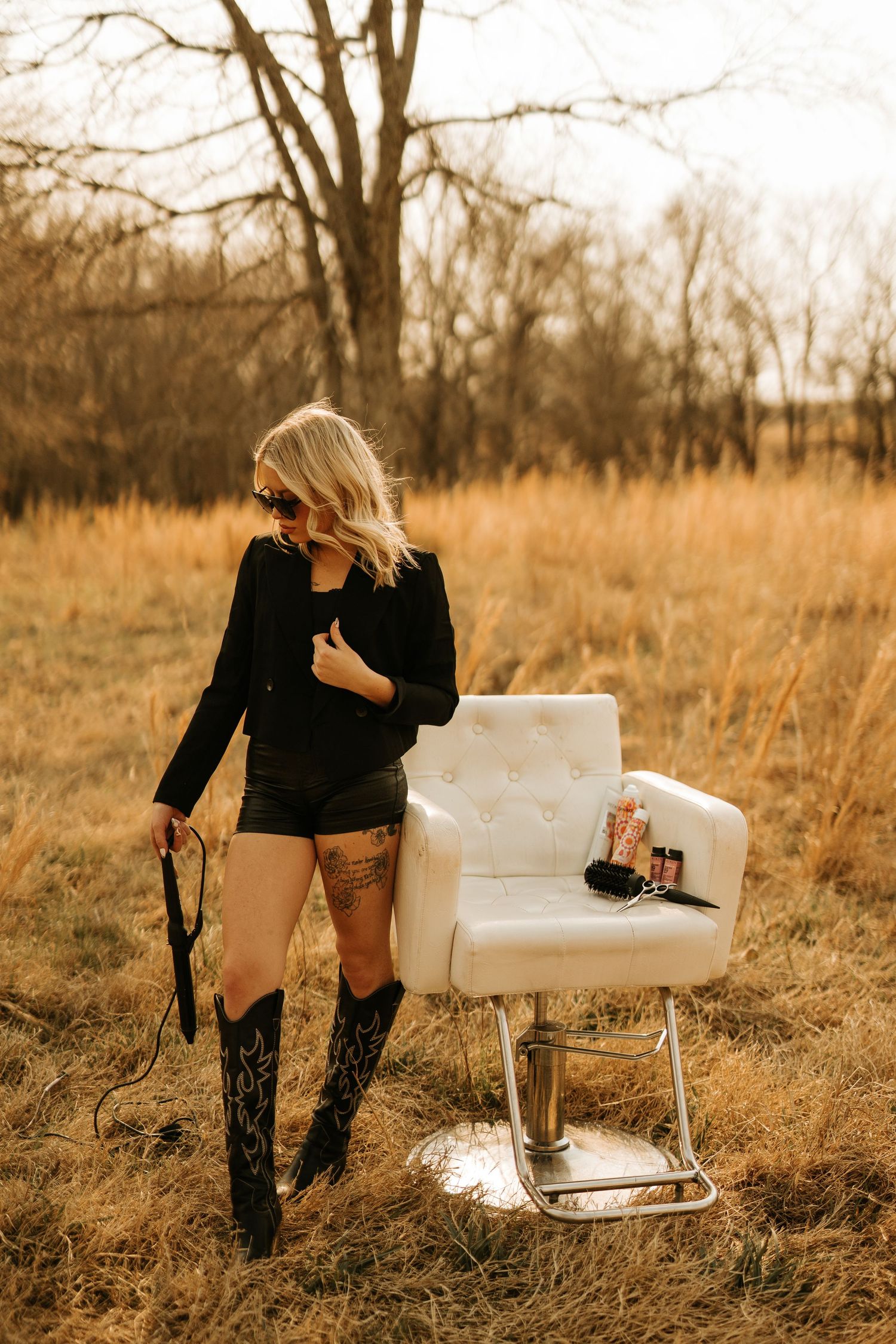 Blonde woman in black outfit and cowboy boots poses outdoors beside a white tufted salon chair in a golden field.