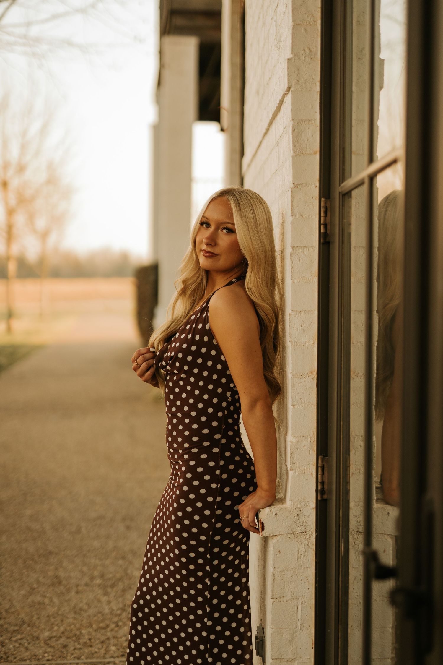 Blonde woman in brown polka dot dress leaning against white brick wall outdoors at golden hour.