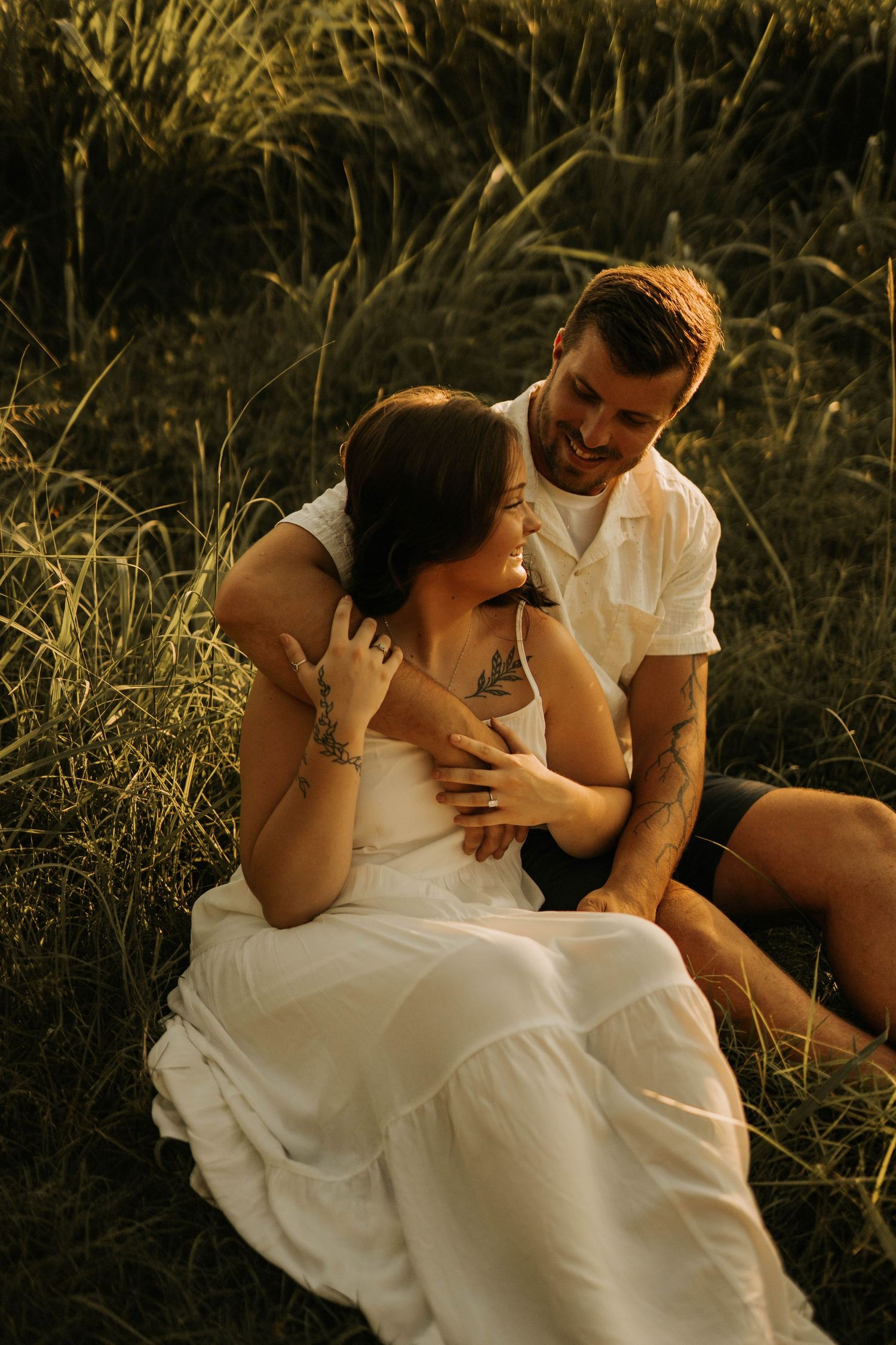 A couple sits in golden grass, smiling and embracing, the woman in a white dress and the man in a white shirt.