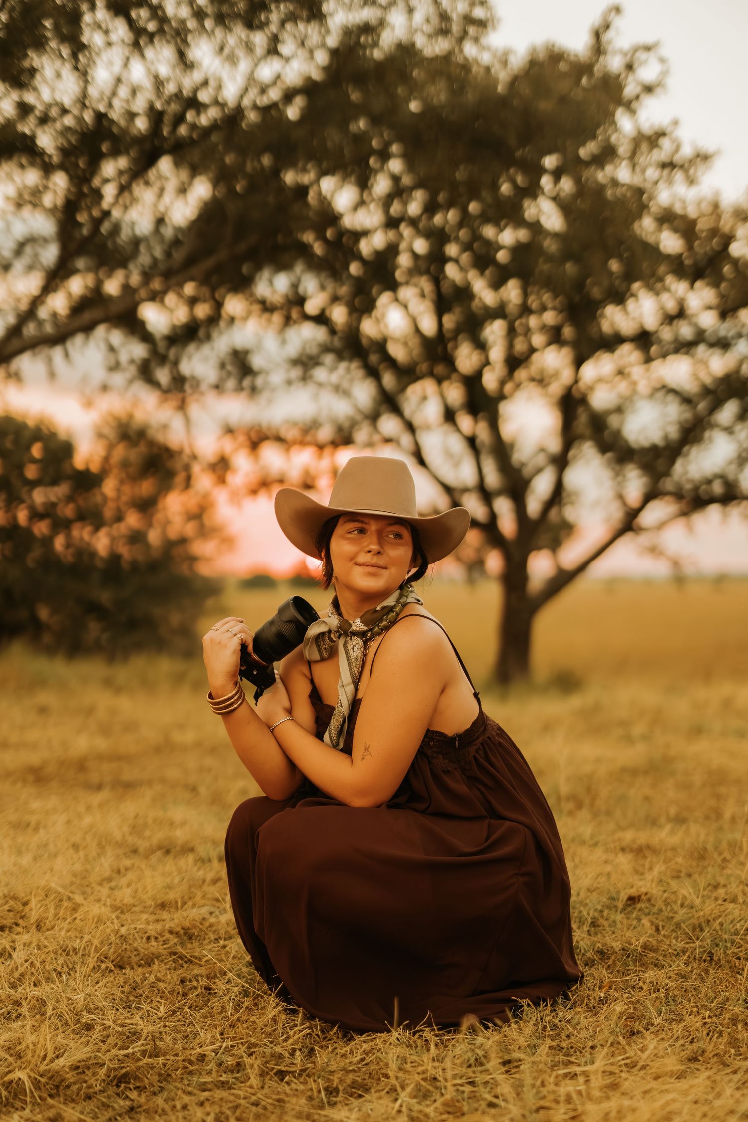 Woman in cowboy hat and dark skirt crouching in golden field at sunset, holding camera near trees.