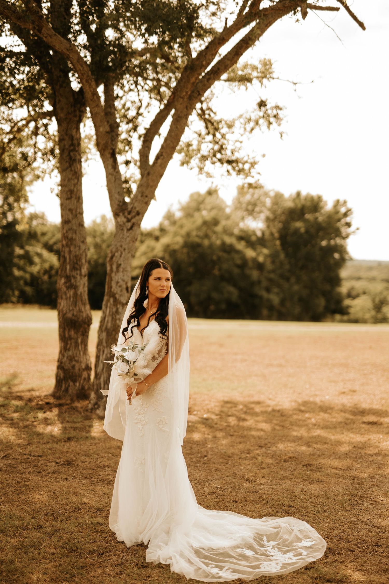 A bride in a flowing white gown poses in a sunlit field with trees.