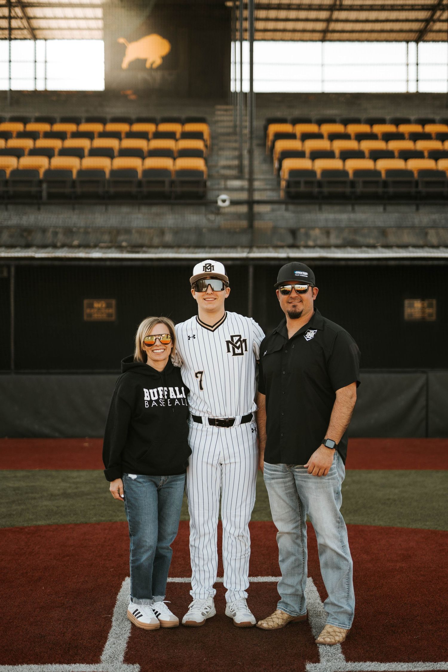 Baseball player poses with parents on field with stadium seating visible in background.