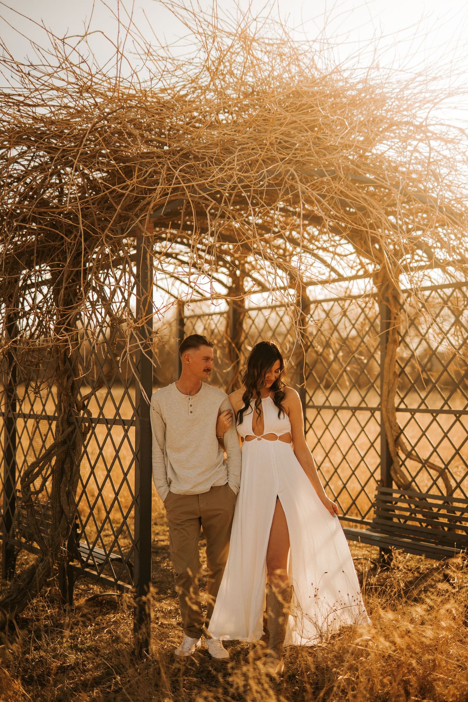 Couple posing romantically under a rustic dried vine arbor in golden sunlight, woman in white dress, man in casual attire.