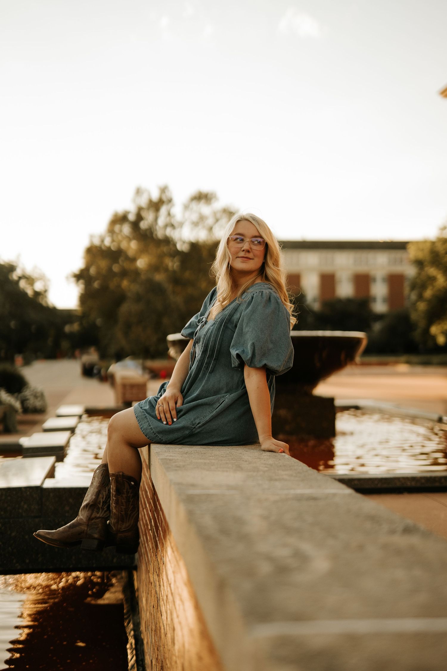 Blonde woman in teal dress and cowboy boots sitting on stone wall near fountain at golden hour.