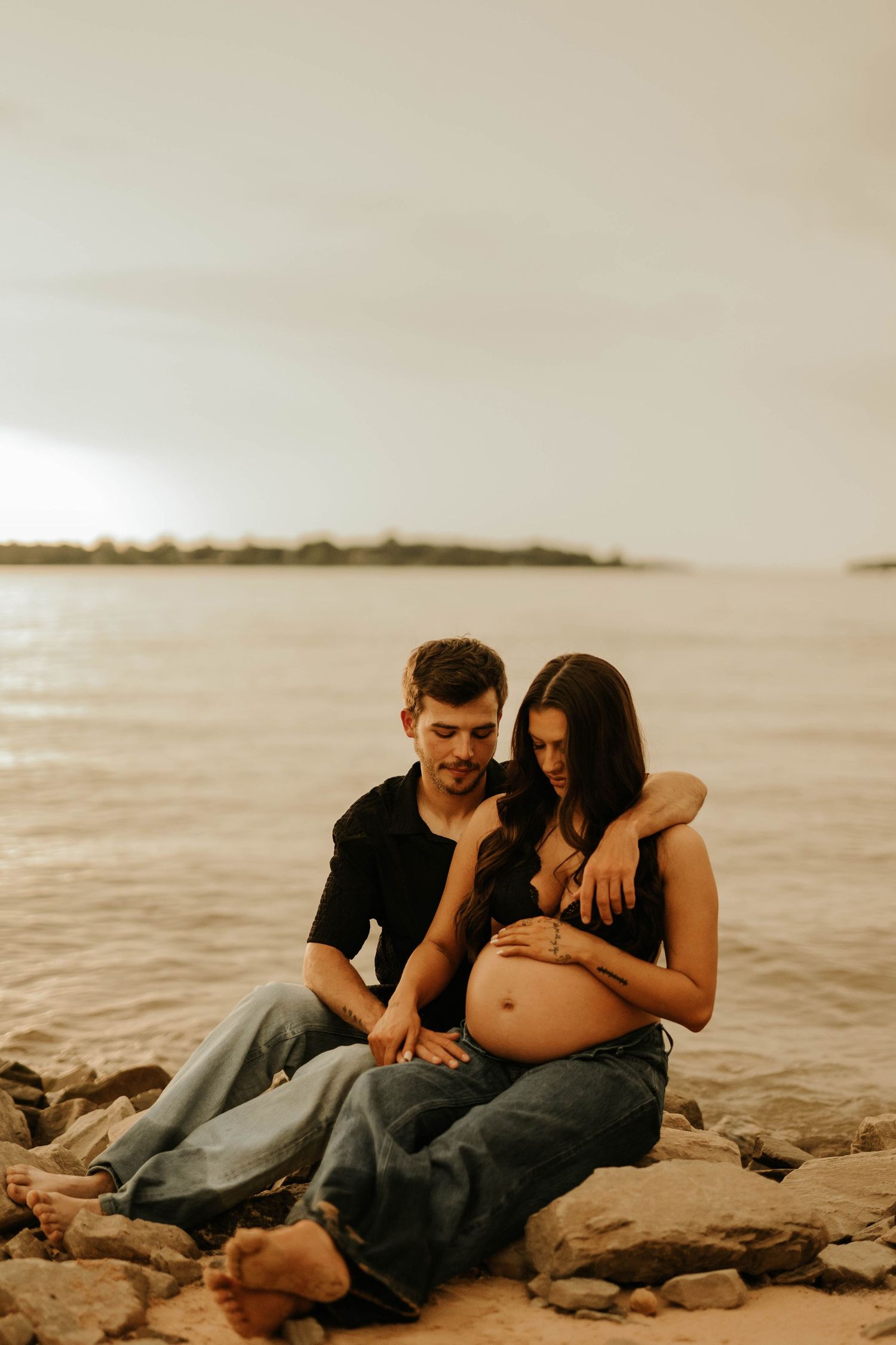 Couple sitting on rocky beach at sunset, man embracing pregnant woman as they look down at her baby bump.