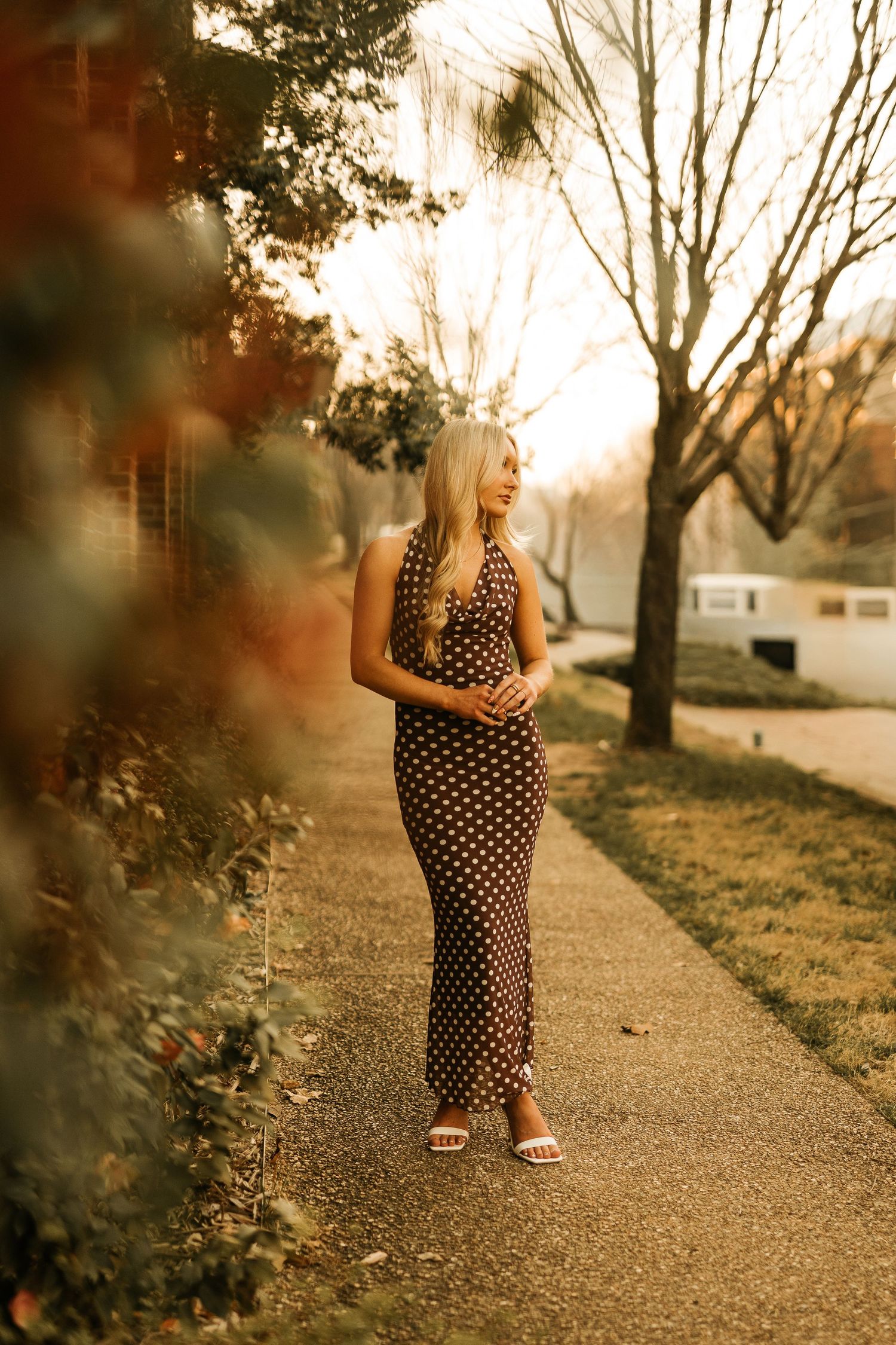 Pregnant woman in polka dot maxi dress standing on sunlit sidewalk surrounded by autumn trees.