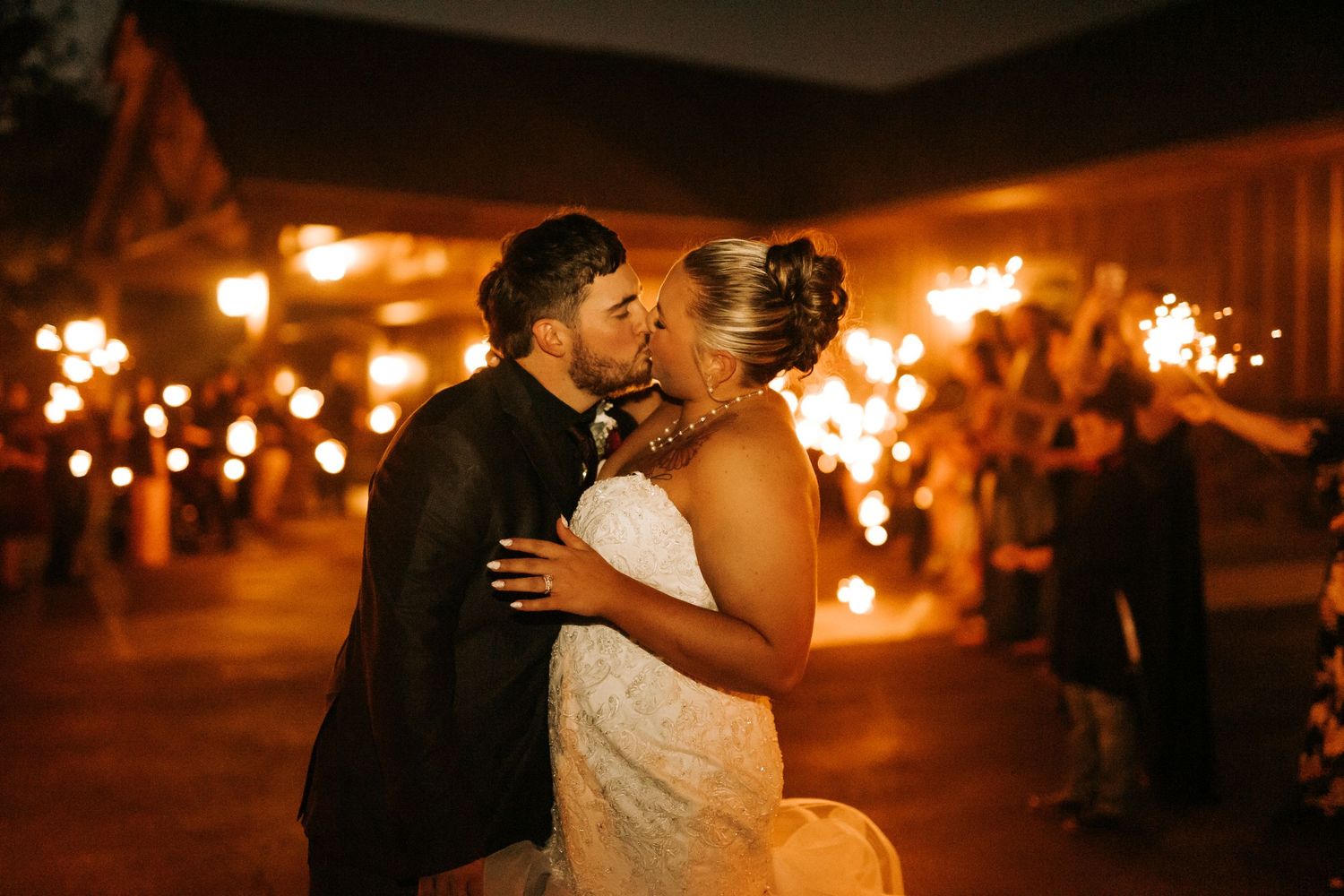 Bride and groom share a romantic kiss at night surrounded by glowing sparklers at their outdoor wedding reception.