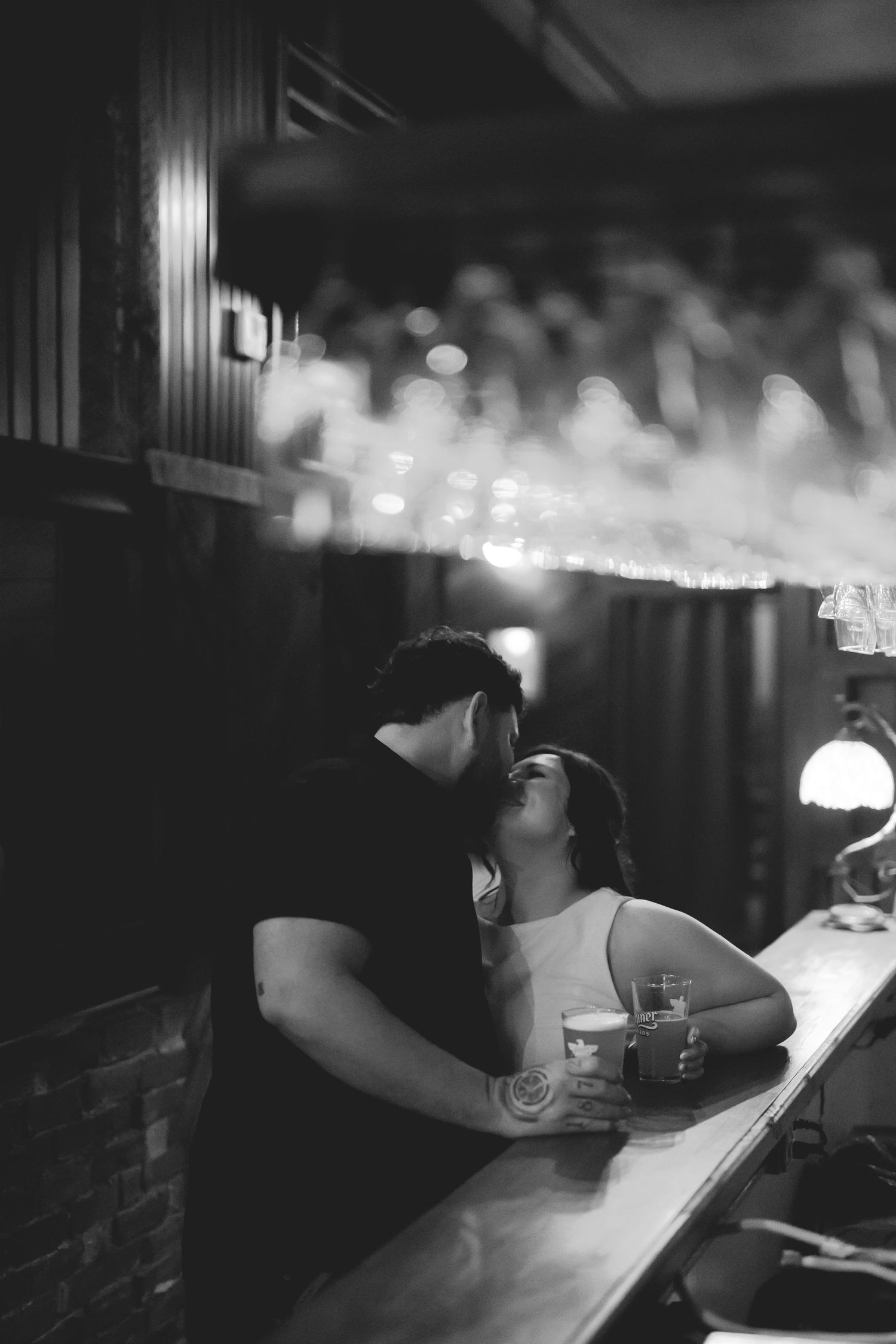 Black and white photo of a couple sharing a romantic kiss at a dimly lit bar counter with drinks nearby.