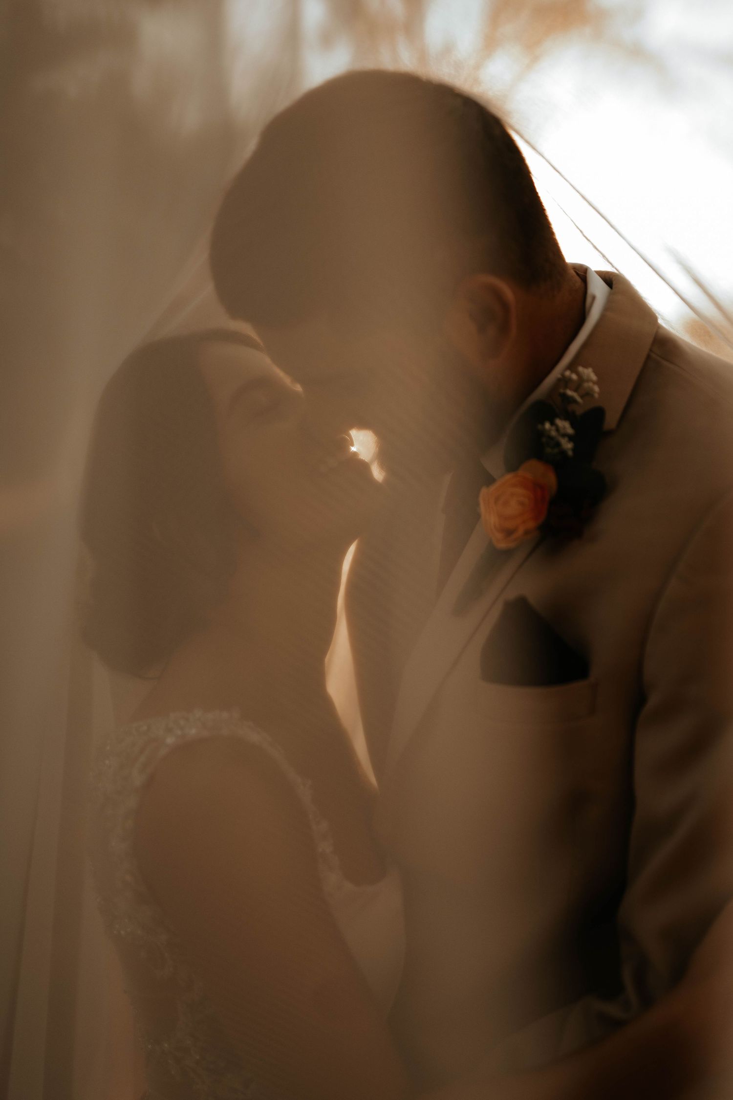 A bride and groom share a romantic moment under a veil, bathed in warm golden light at their wedding.