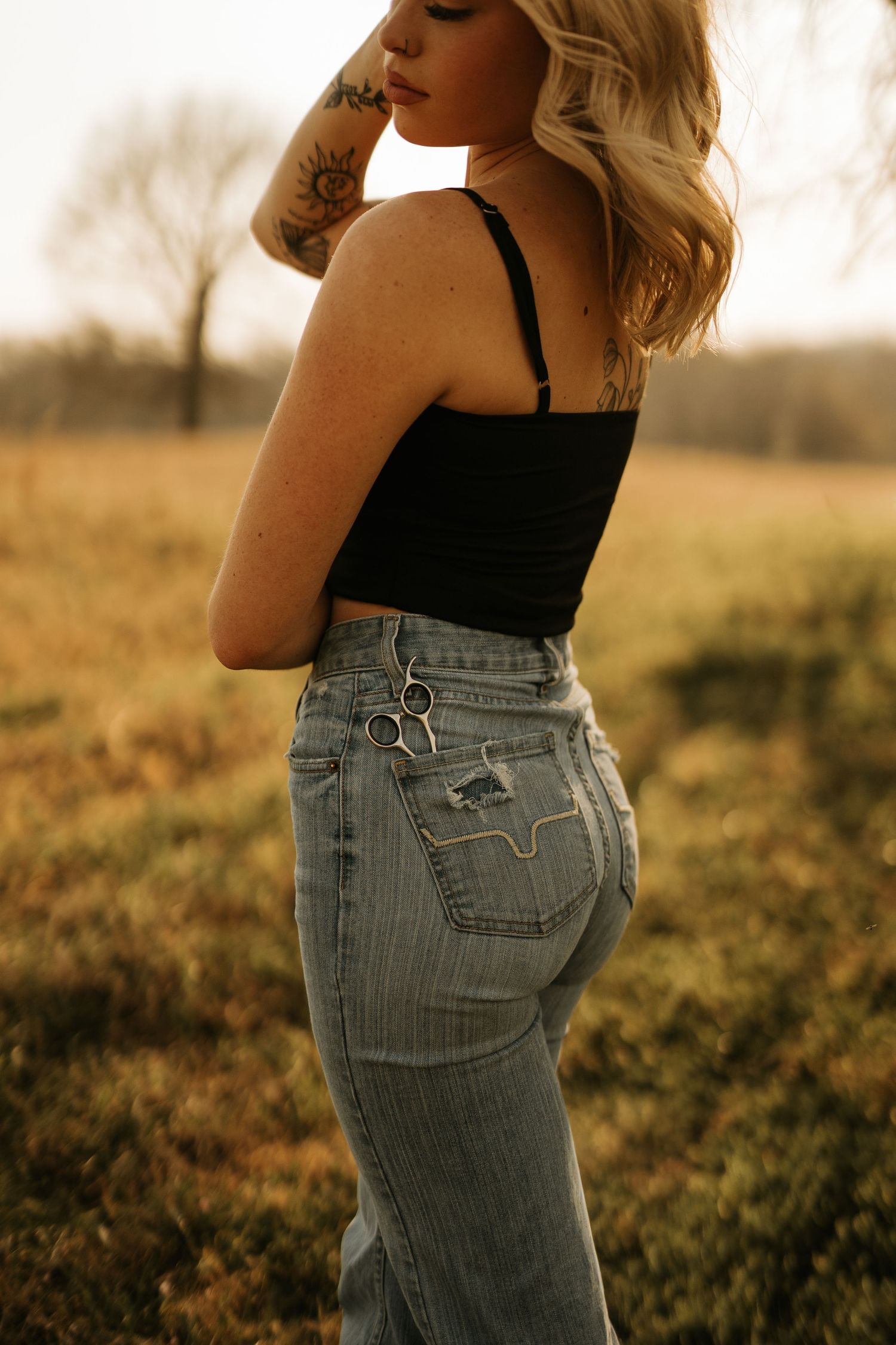 Blonde woman with tattoos wearing black crop top and light wash high-waist jeans with scissors in pocket, standing in field.