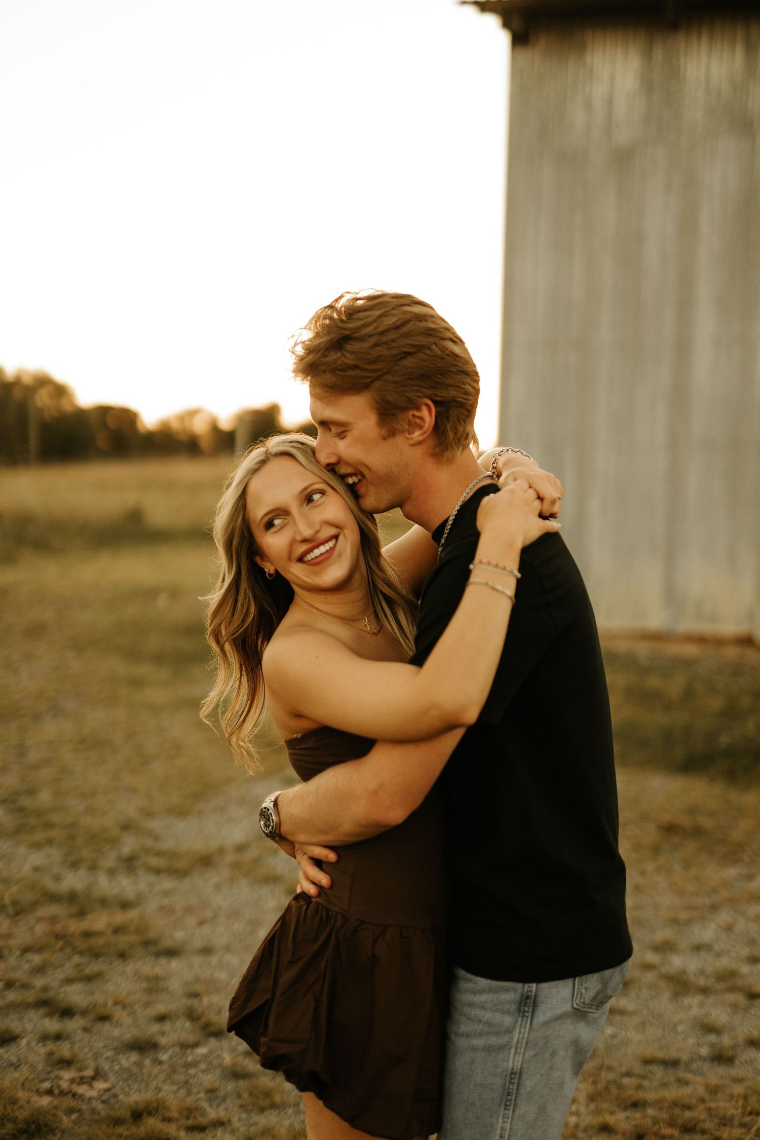 A smiling couple embraces outdoors at golden hour near a rustic barn, the woman in a brown dress hugging a man in a black shirt.