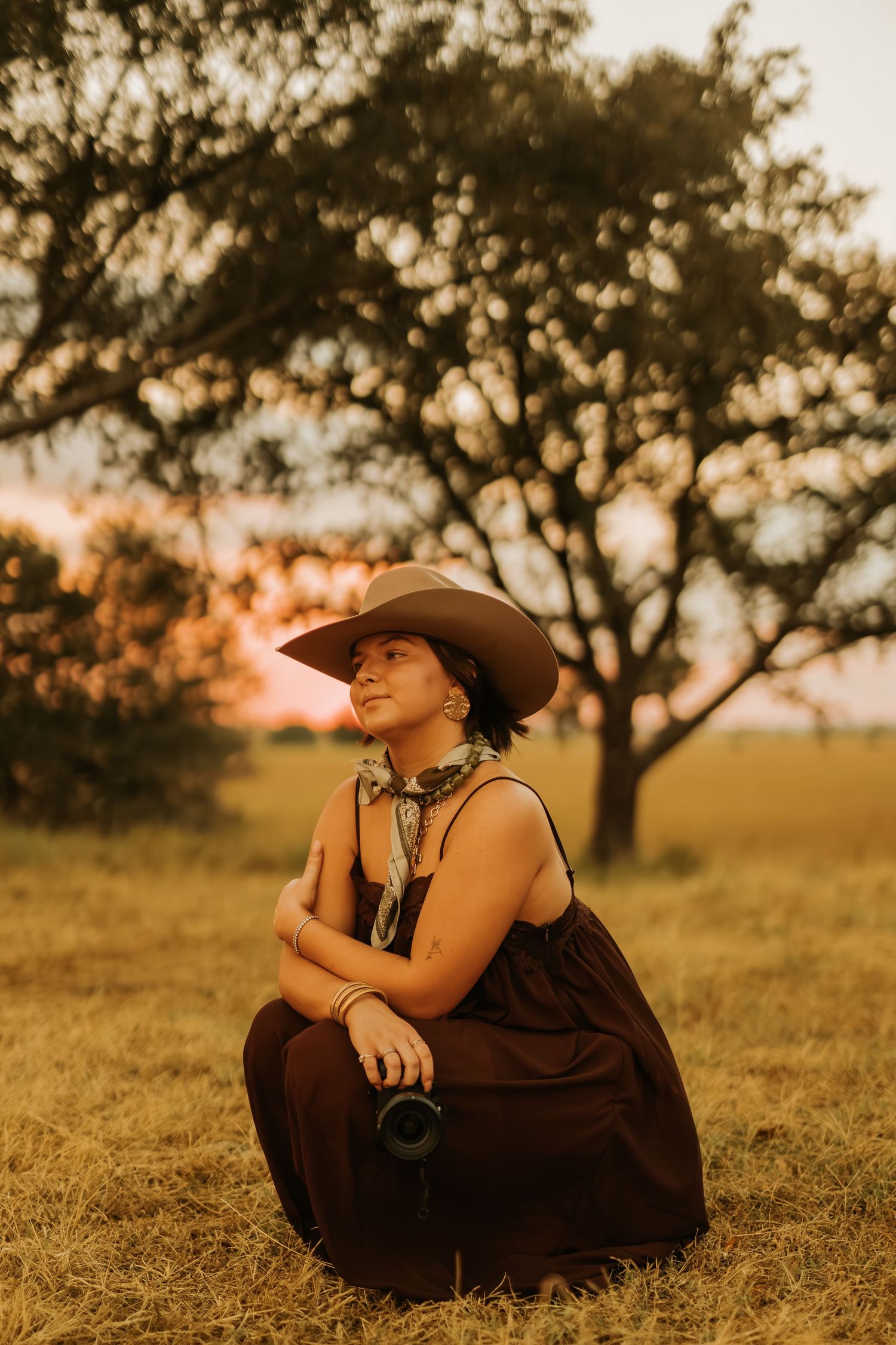 Woman in wide-brim hat and dark dress sits in golden field at sunset, holding camera near a tree.