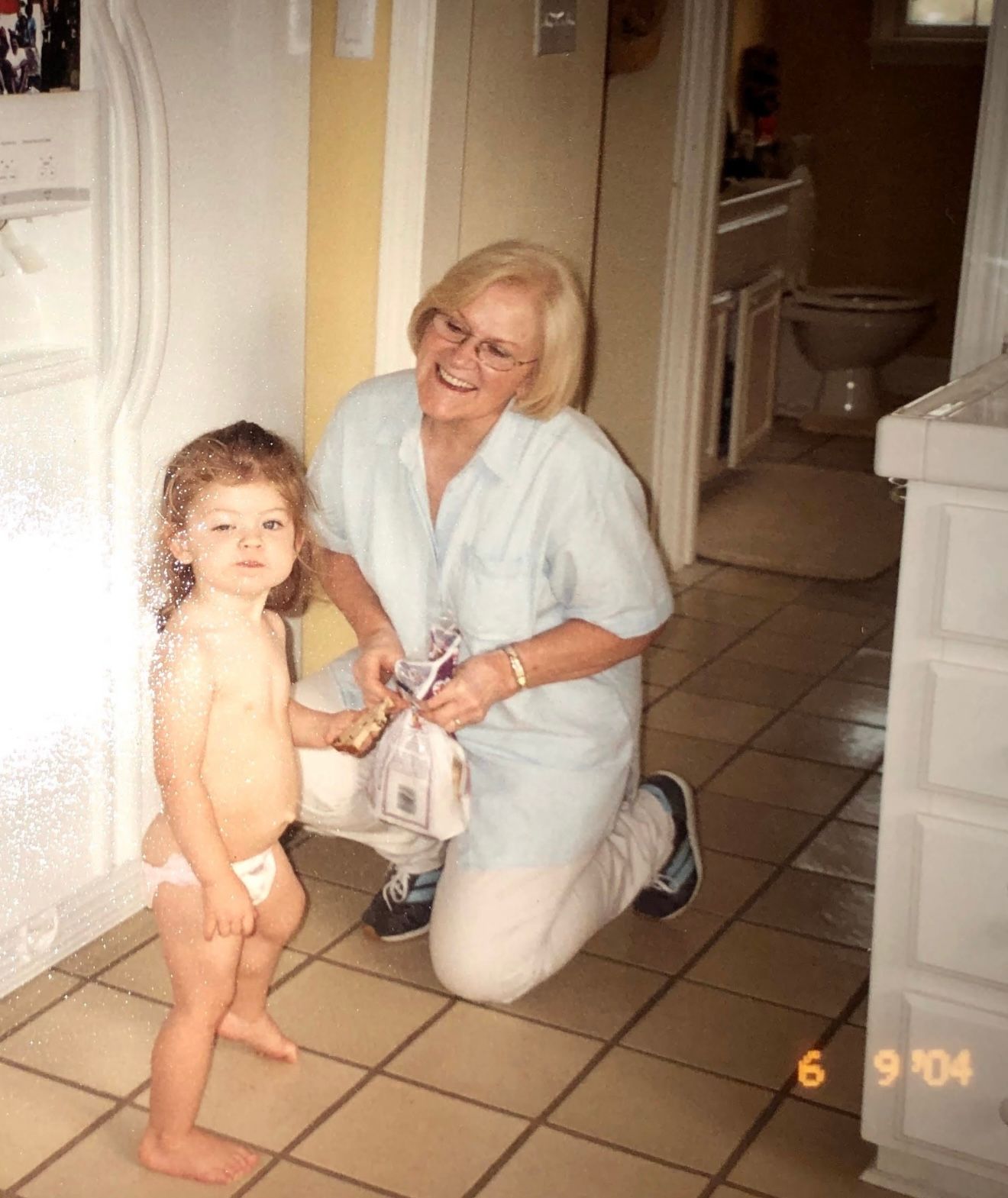 Elderly woman kneeling beside a toddler in diapers on a tiled floor, holding a gift bag, indoors.