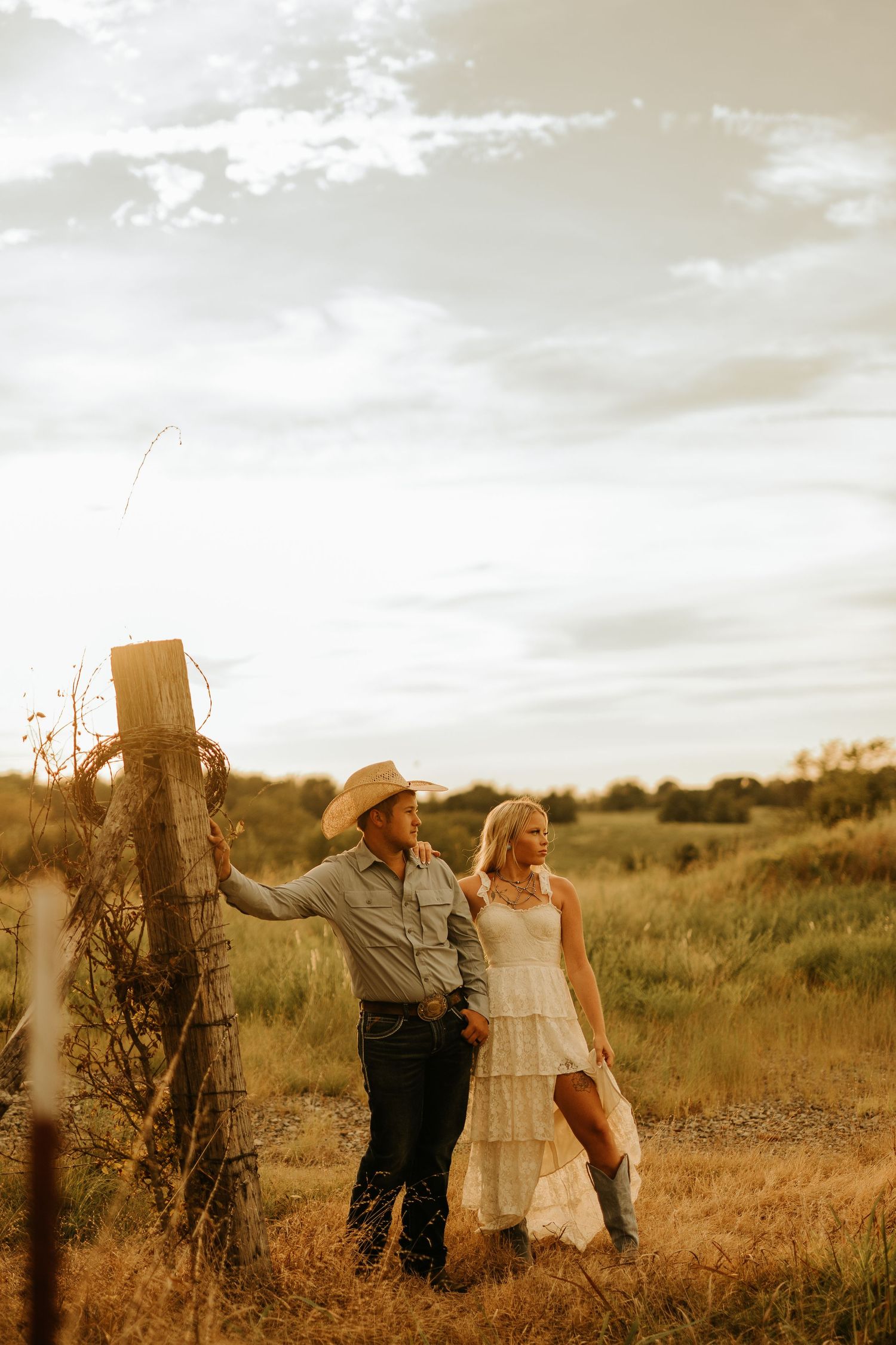 A couple in western attire stands by a fence post in a golden field at sunset, man in cowboy hat, woman in white lace dress.