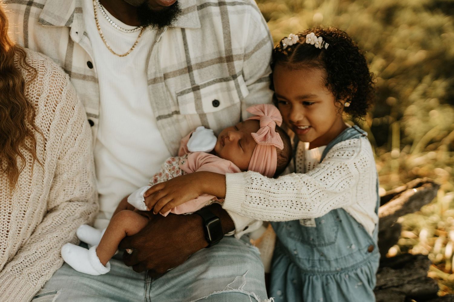 A father holds a newborn baby in pink while a young girl in denim overalls gently touches the infant outdoors.