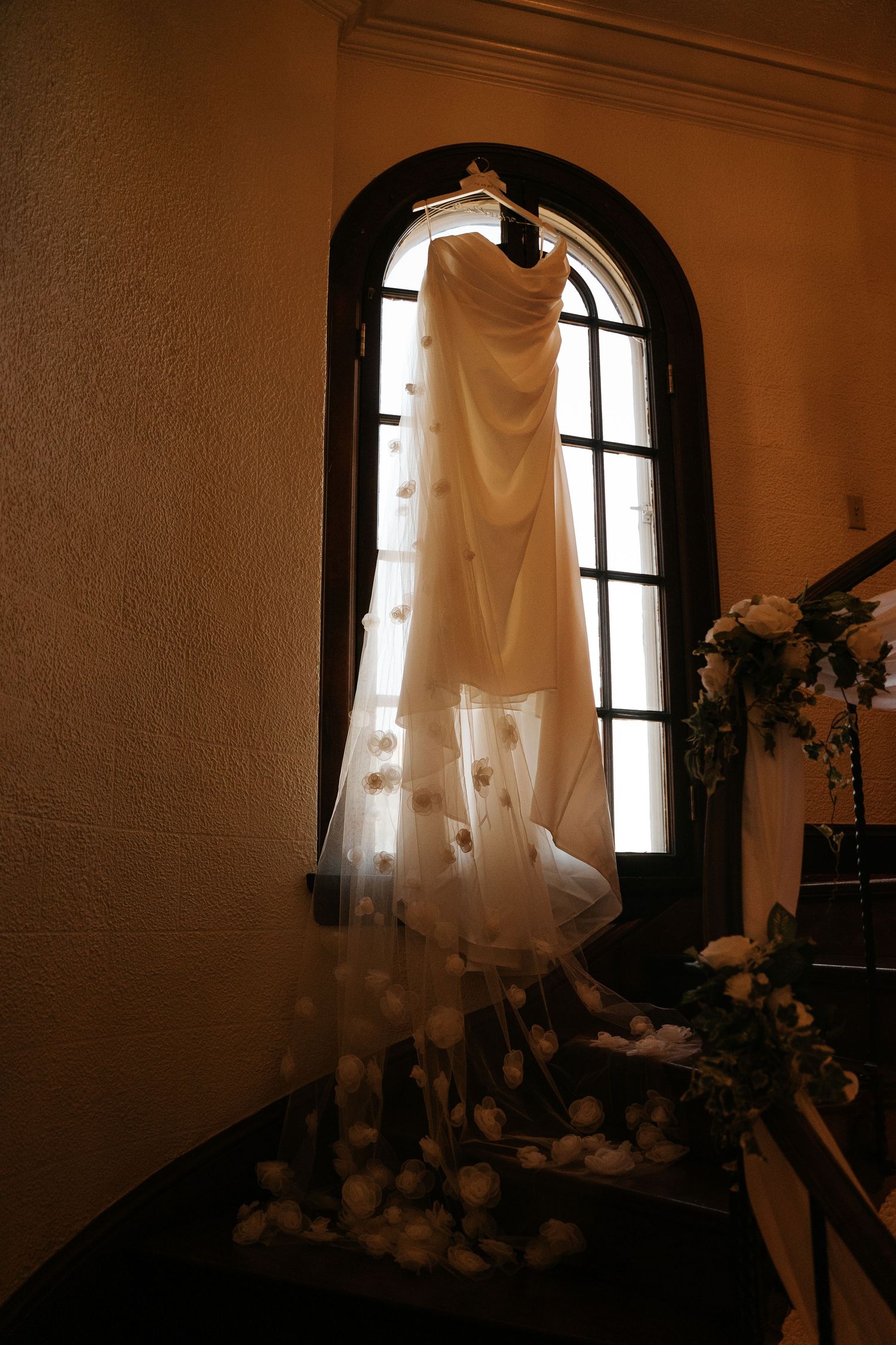 Wedding dress hanging in arched window with floral details cascading down staircase railing in warm elegant venue.