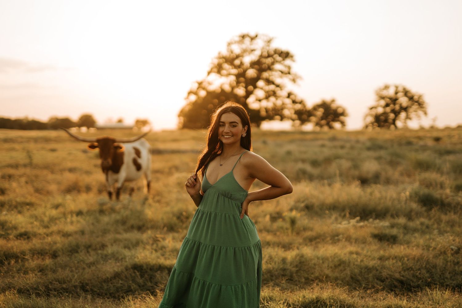 Woman in green dress poses at golden hour in a field with a longhorn cow grazing in the background.