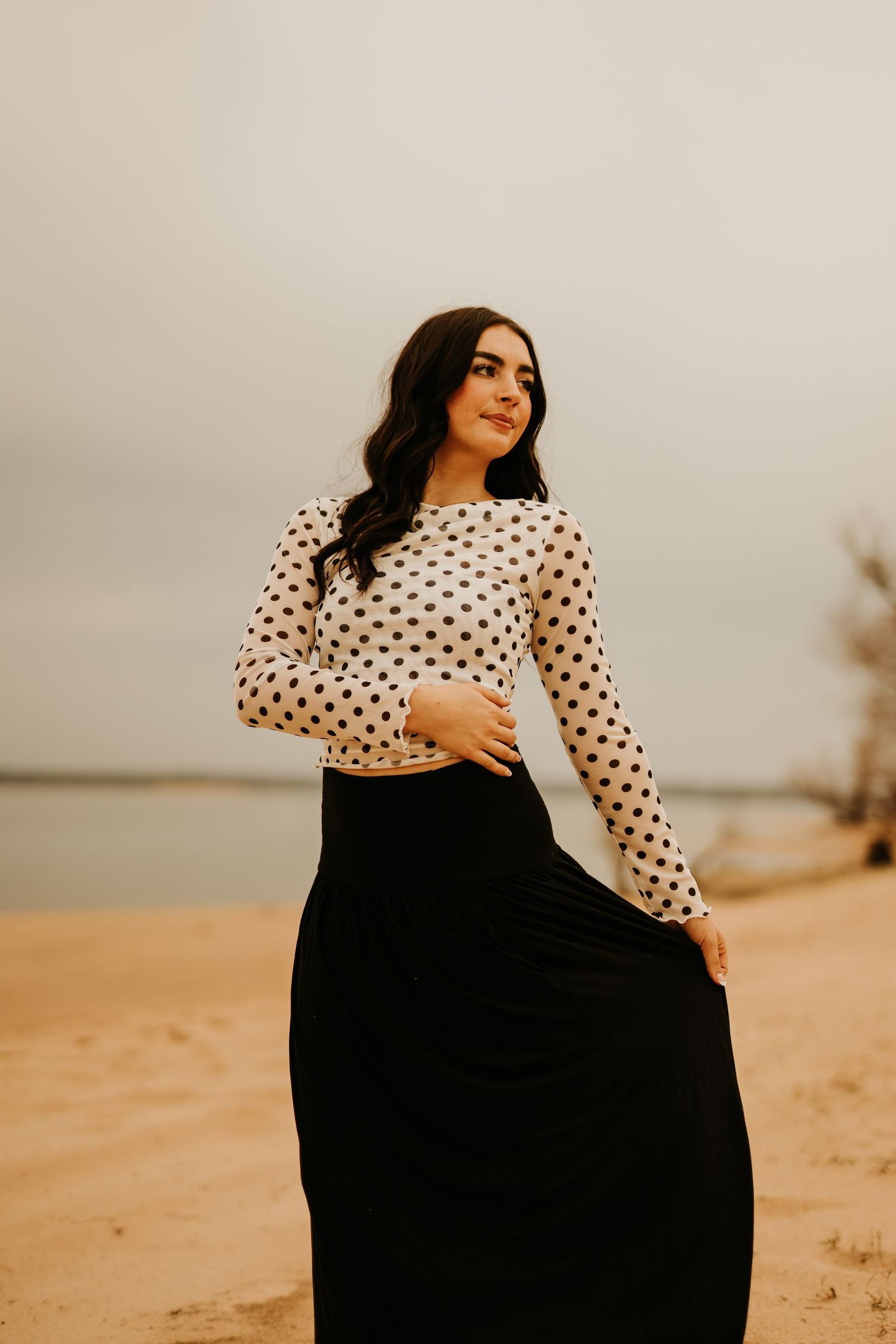 Woman in polka dot crop top and black maxi skirt posing on sandy beach with overcast sky.