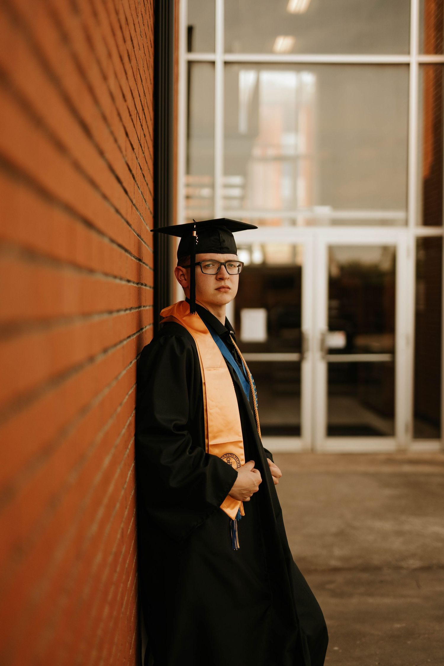 Graduate in traditional academic regalia poses against brick wall in moody lighting.