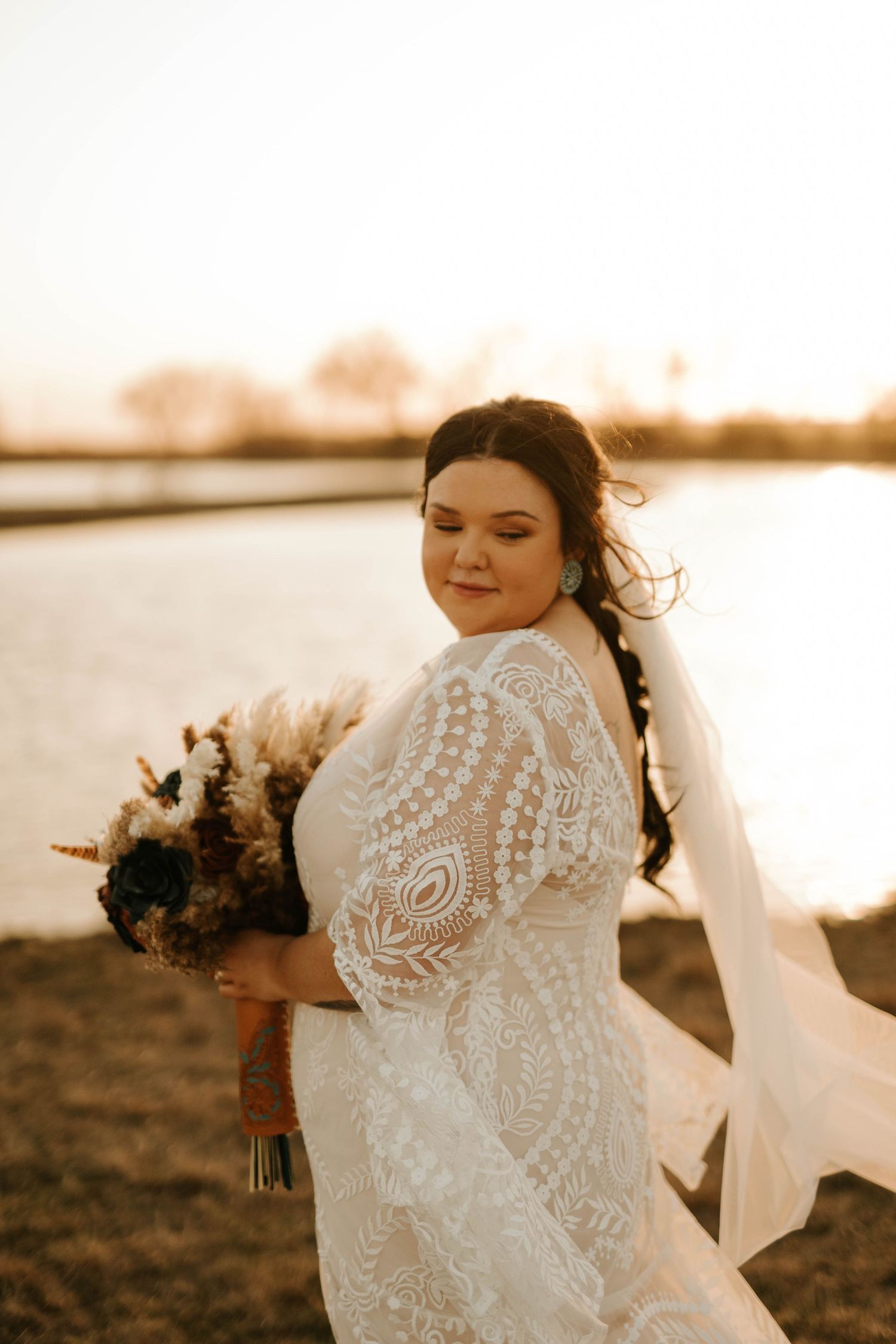 Bride in detailed lace wedding dress poses at sunset by a lake holding a rustic bouquet.