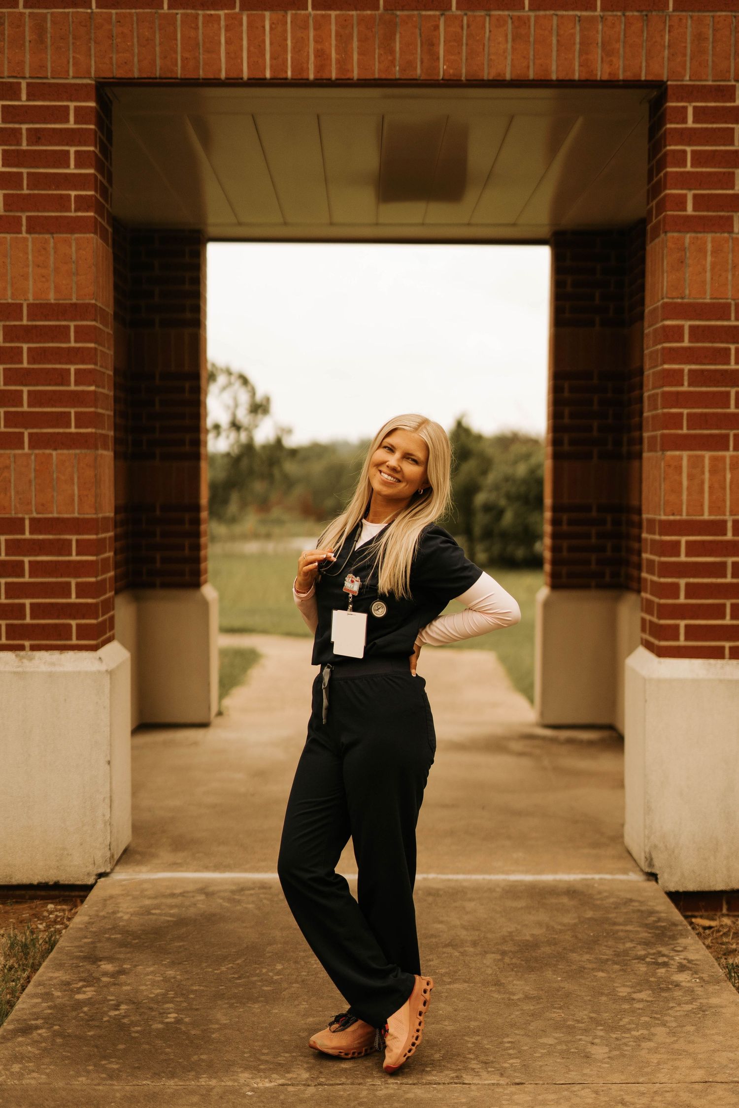 Healthcare worker in black scrubs poses confidently in brick archway during golden hour.