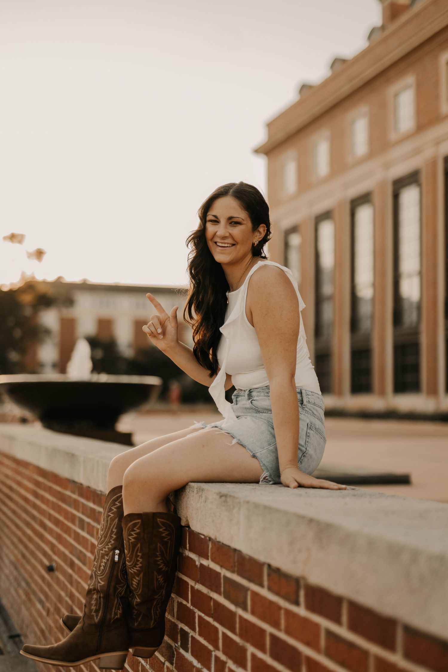 A person in western boots sits on a brick ledge during sunset wearing casual summer attire.