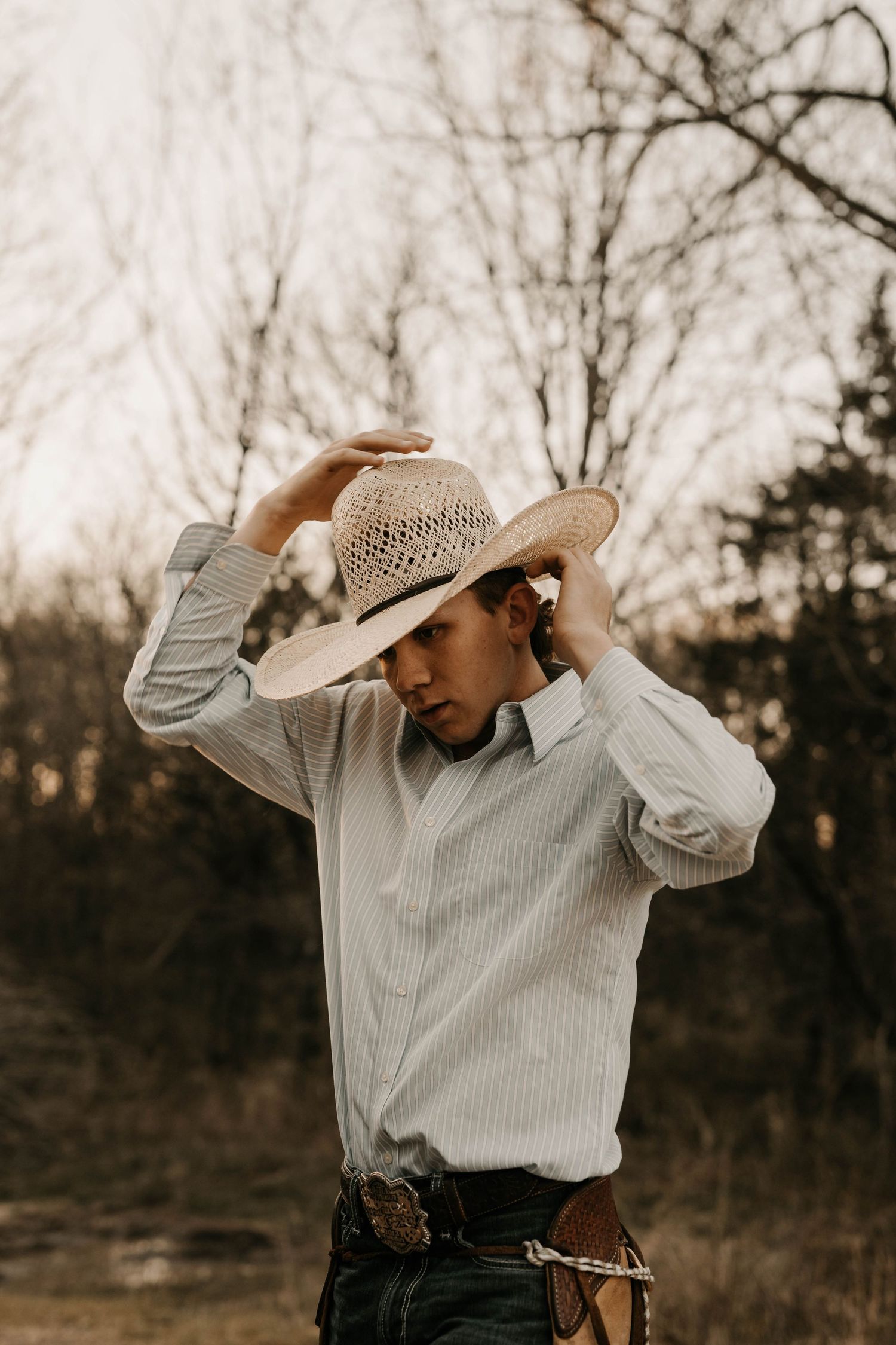 Person adjusting cowboy hat while wearing western-style clothing outdoors.