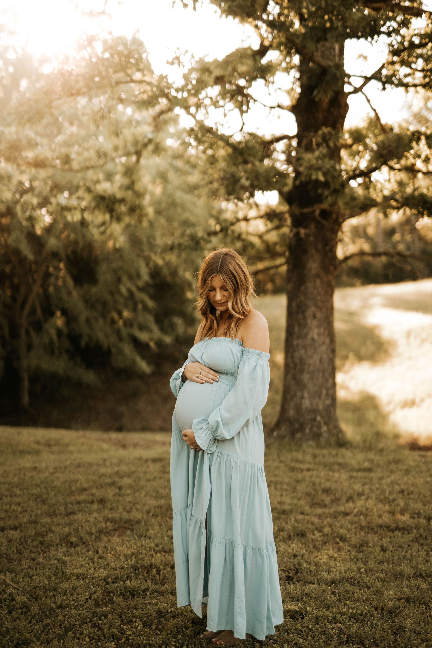 Woman in flowing light blue dress poses in garden during golden hour maternity photoshoot.