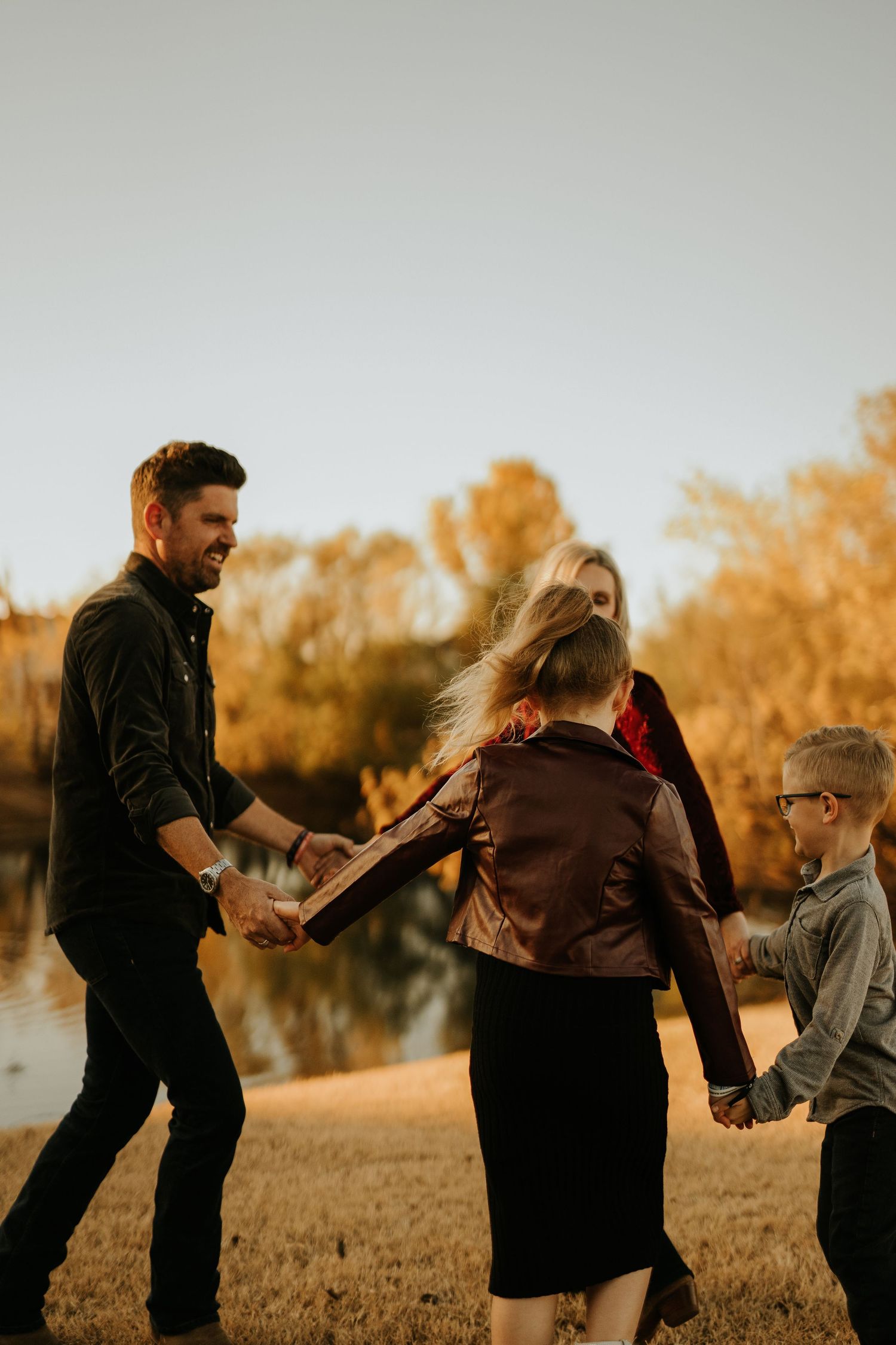 Family holding hands and playing together outdoors near a pond during golden autumn sunset.