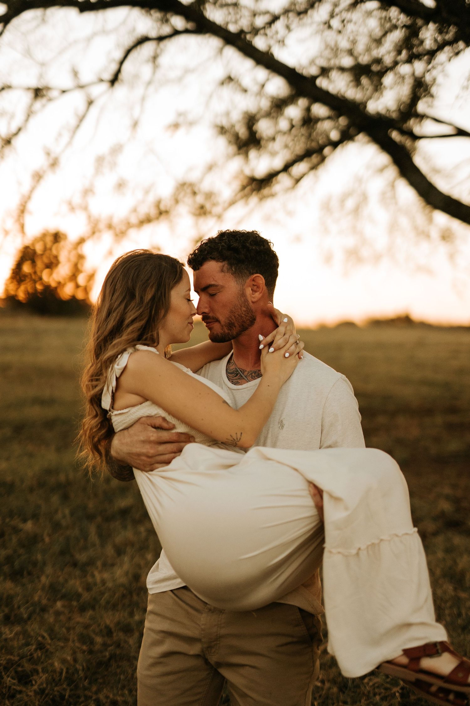 A man lifts a woman in a white dress as they share a romantic moment at golden hour under a tree.
