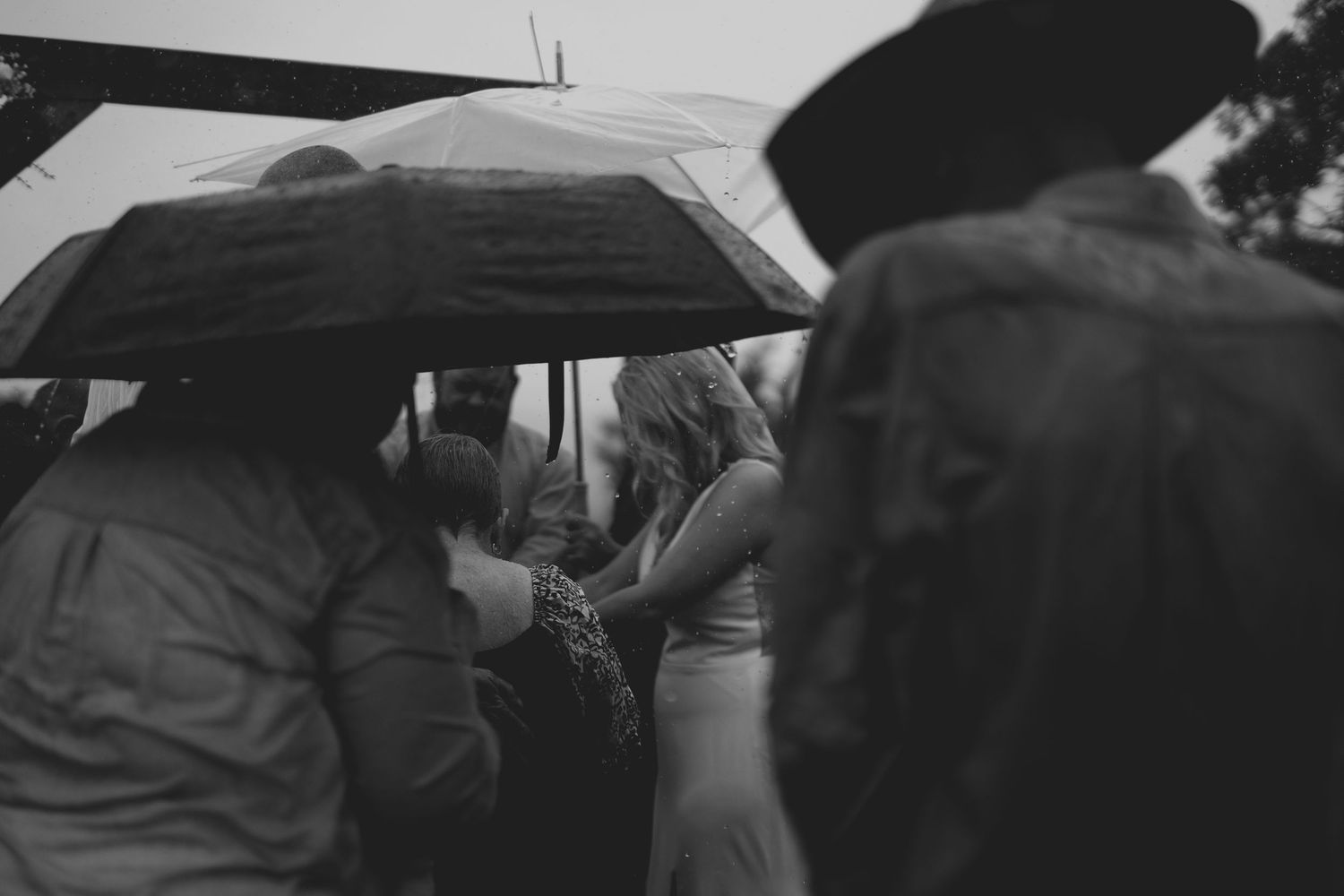 Black and white photo of wedding guests huddled under umbrellas in the rain during an outdoor ceremony.