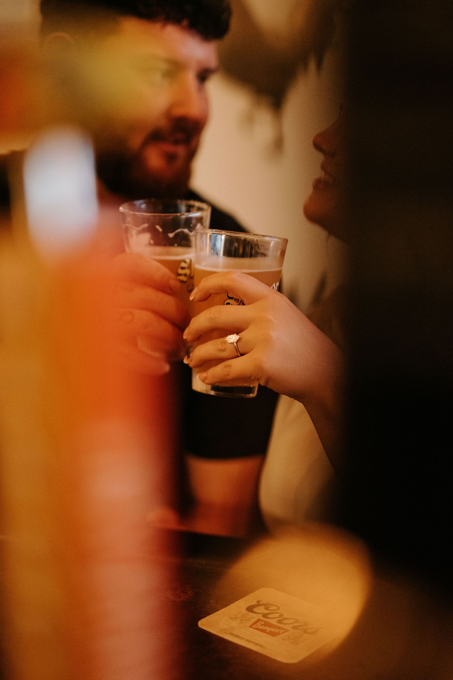 Couple clinking pint glasses of beer at a bar, woman wearing engagement ring, warm bokeh lighting.