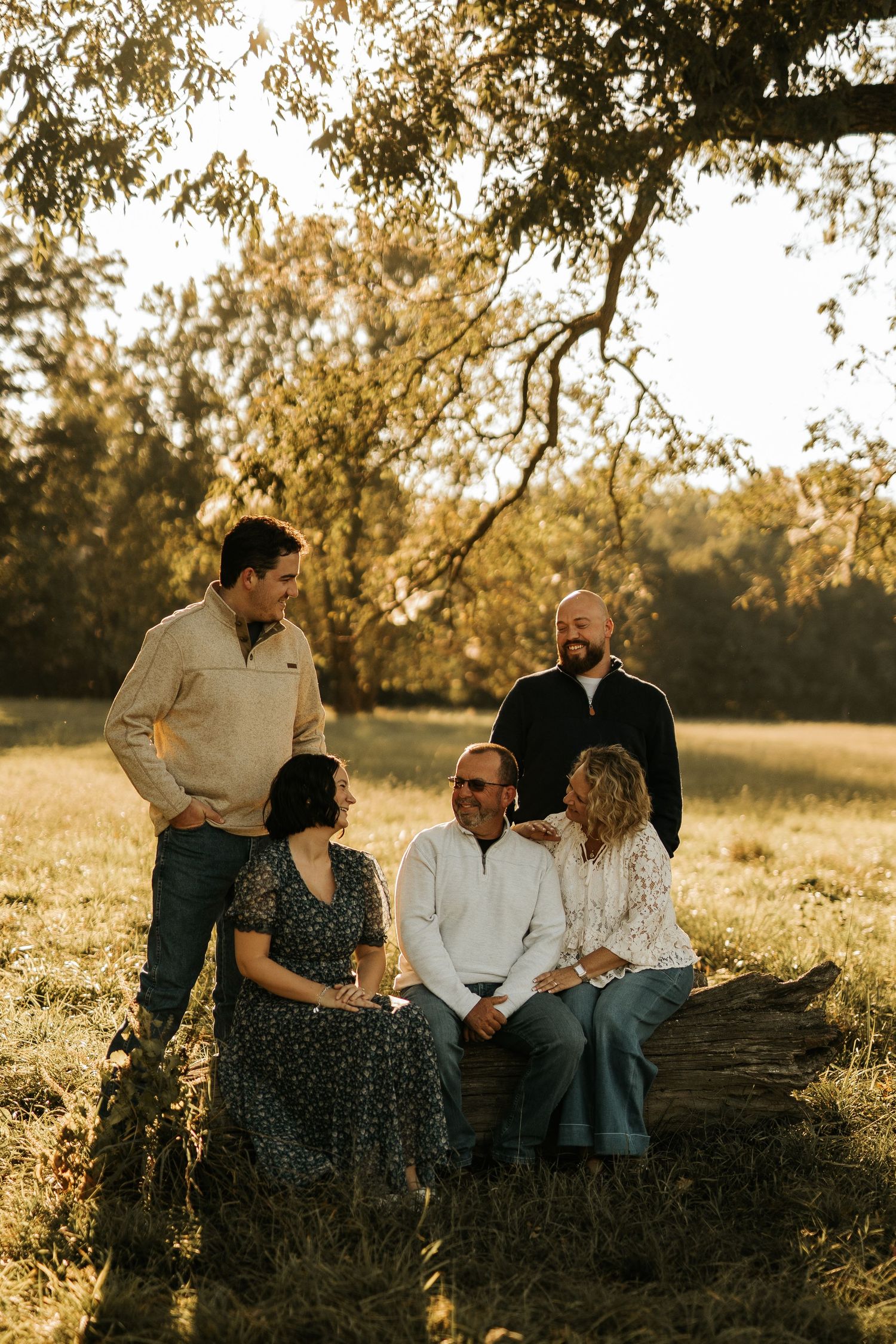 Family of five poses outdoors at golden hour near a fallen log surrounded by lush green trees.