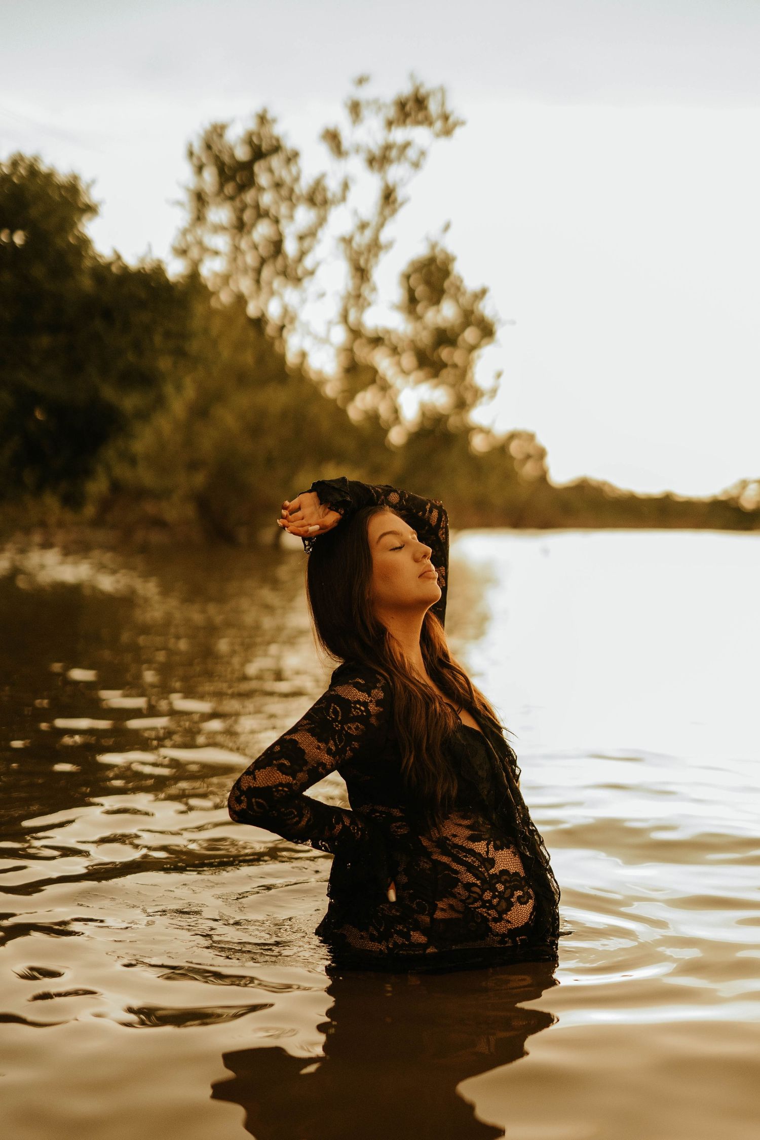 Woman in black lace dress standing in a sunlit river at golden hour, eyes closed, hand in long dark hair.