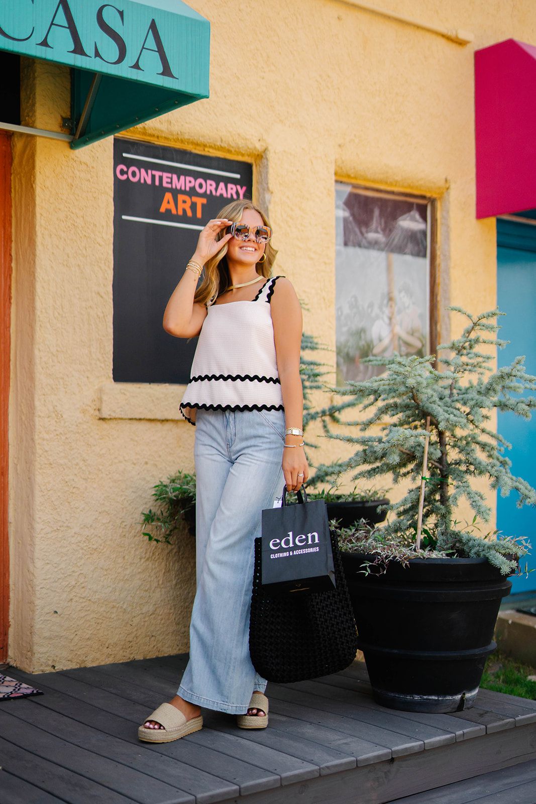 Woman in scalloped tank top and wide-leg jeans stands by contemporary art gallery entrance.