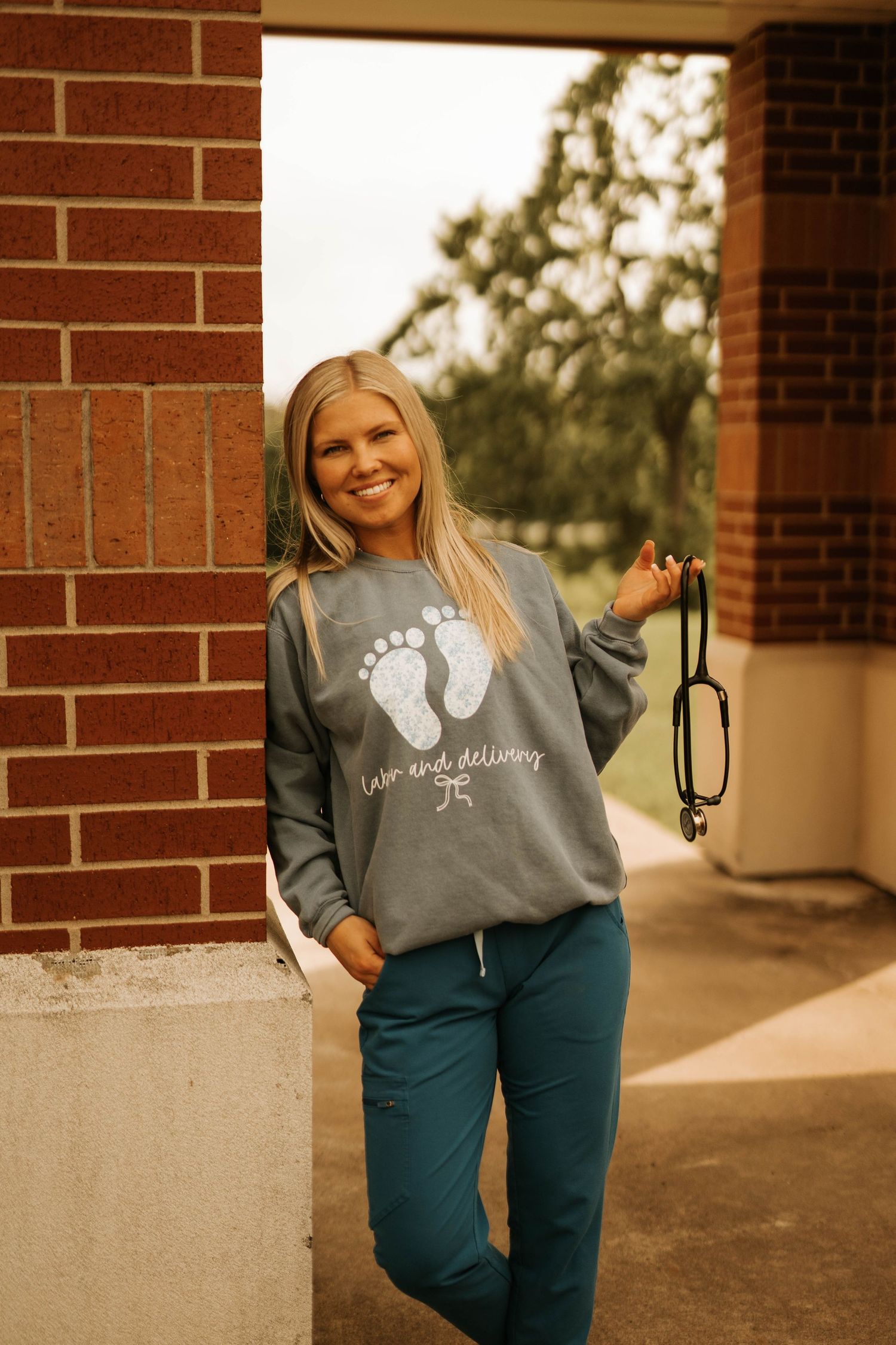Medical professional wearing gray sweatshirt with footprint design stands in brick doorway.