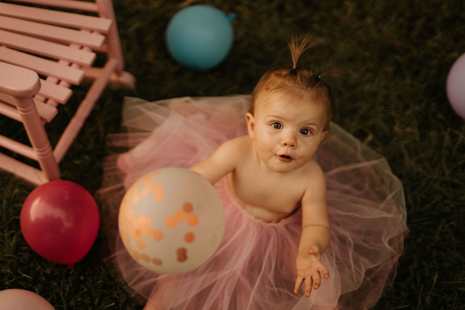 Baby girl in pink tutu sits on grass surrounded by colorful balloons at her first birthday party.