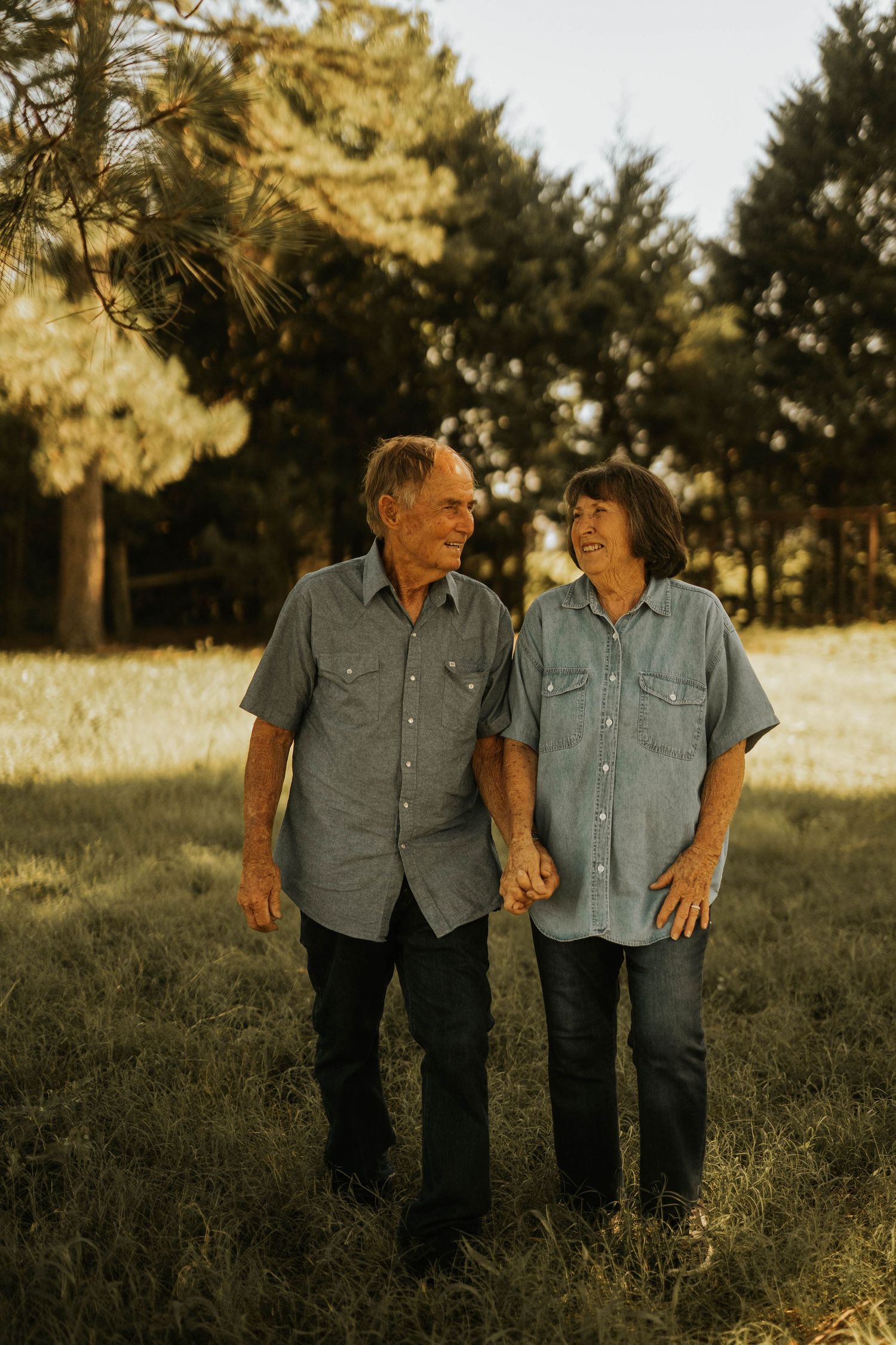 An older couple in matching blue chambray shirts holds hands smiling at each other in a sunlit grassy field.