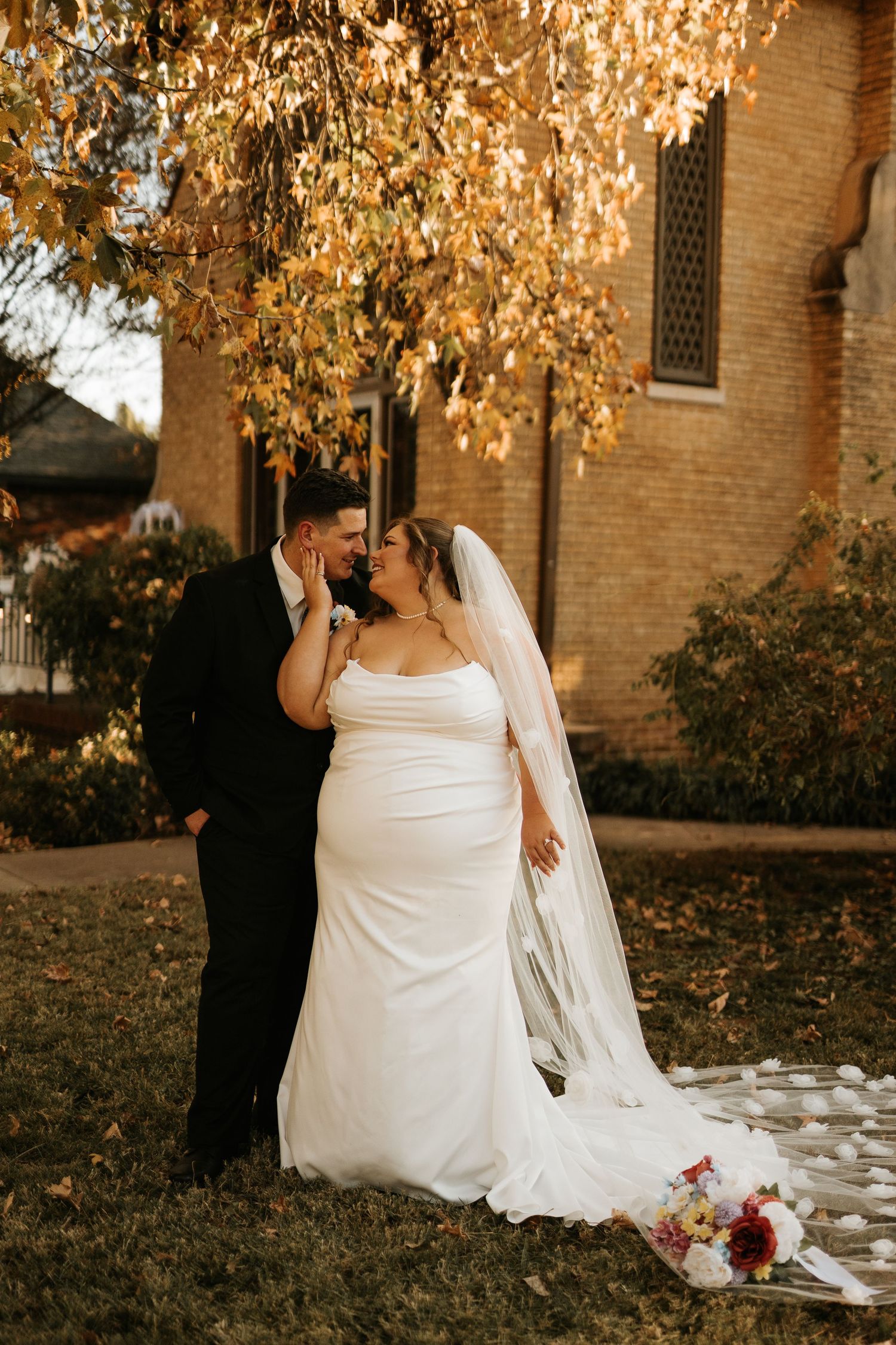Bride and groom share a romantic moment under golden autumn tree beside brick church in fall wedding portrait.
