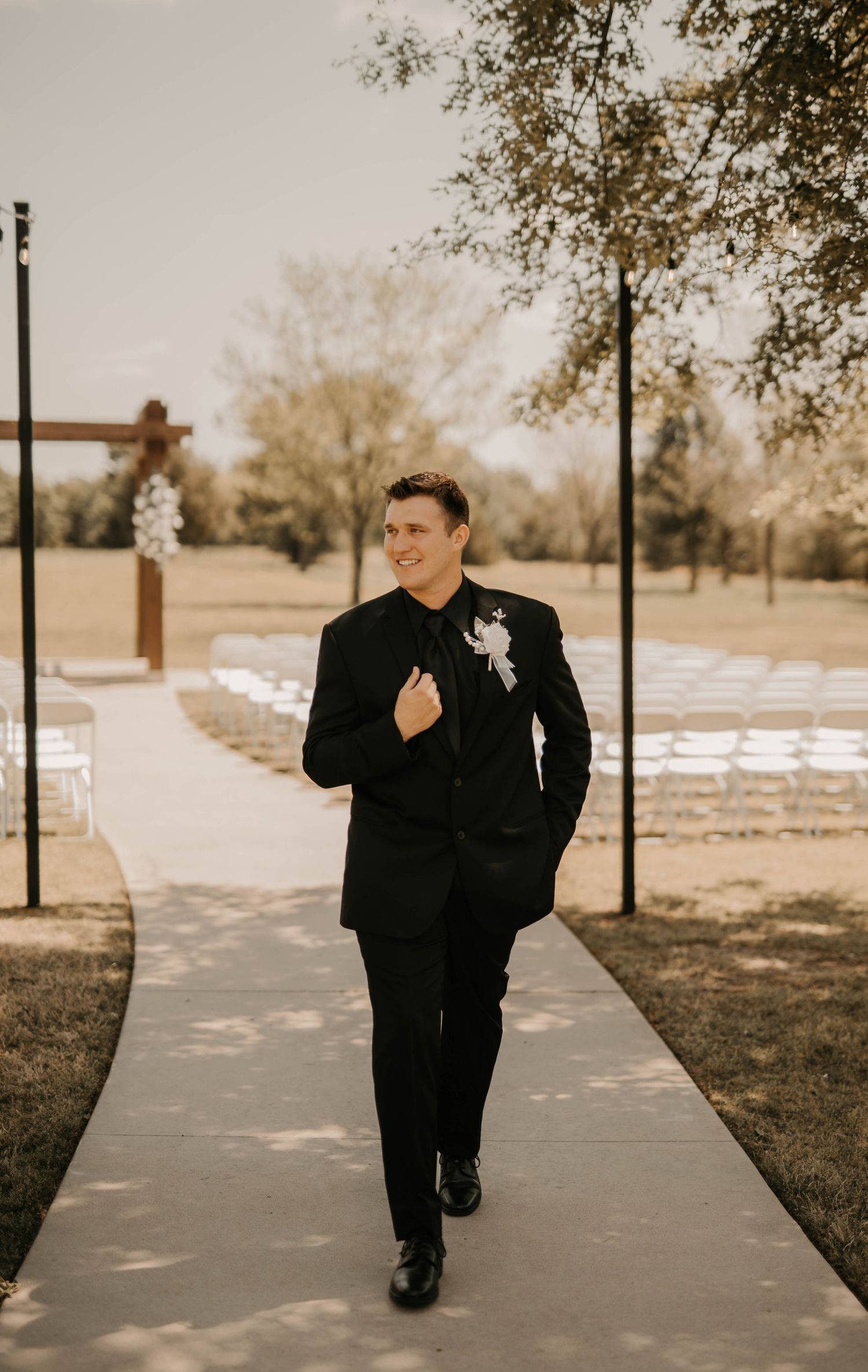 A groom in a classic black suit walks down a pathway with white chairs set up for an outdoor ceremony.