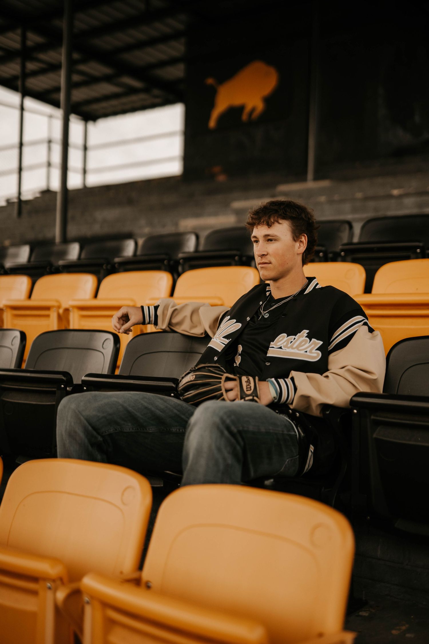 Someone sits alone in stadium seating wearing a varsity jacket while orange seats surround them.
