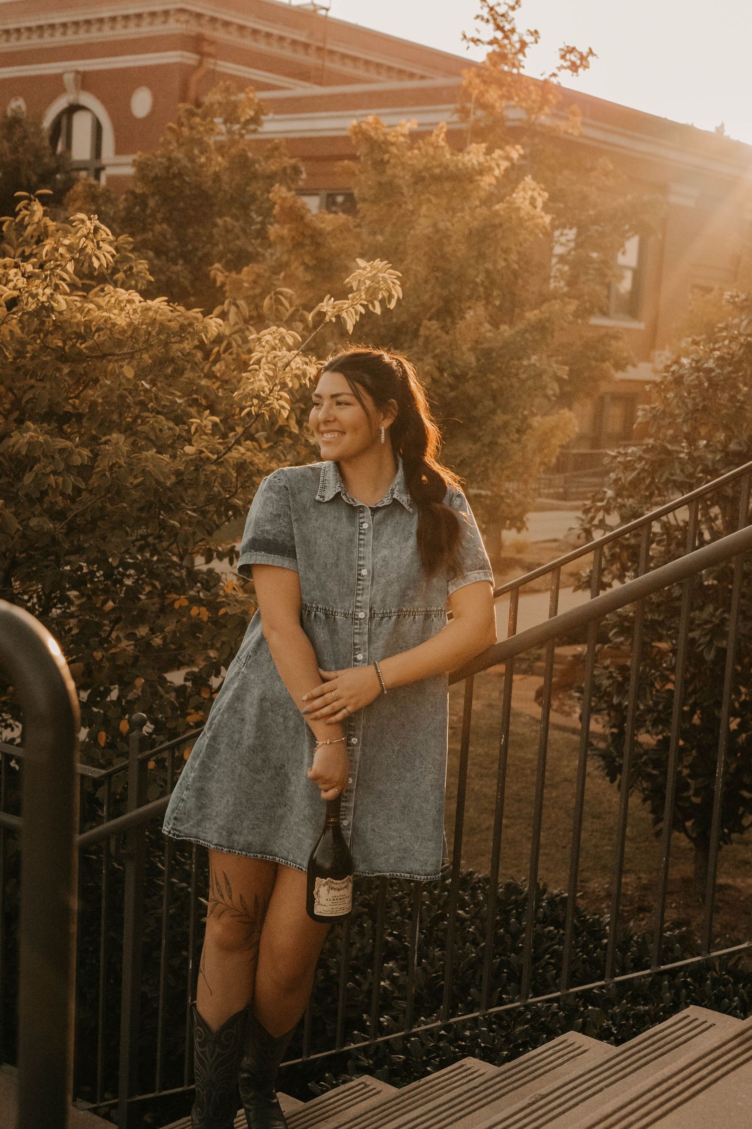 Person in denim dress poses on steps of brick building during sunset.
