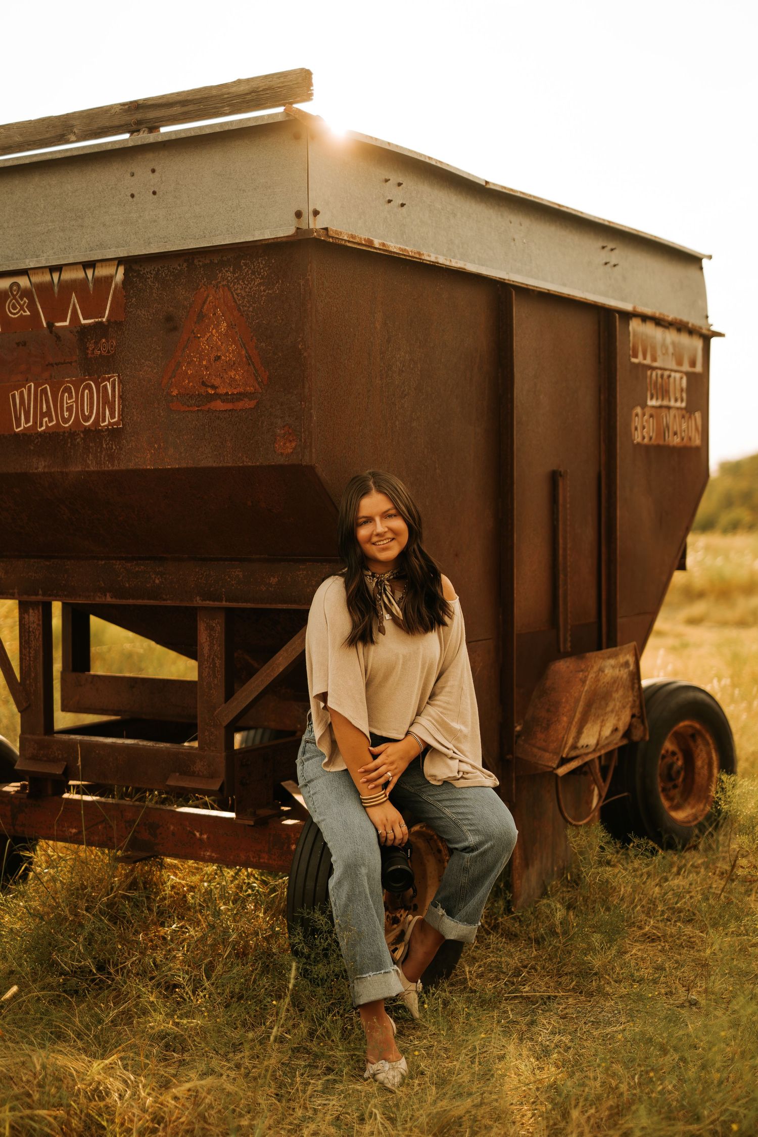 Young woman in jeans sitting on rusty farm wagon at golden hour in a grassy field.