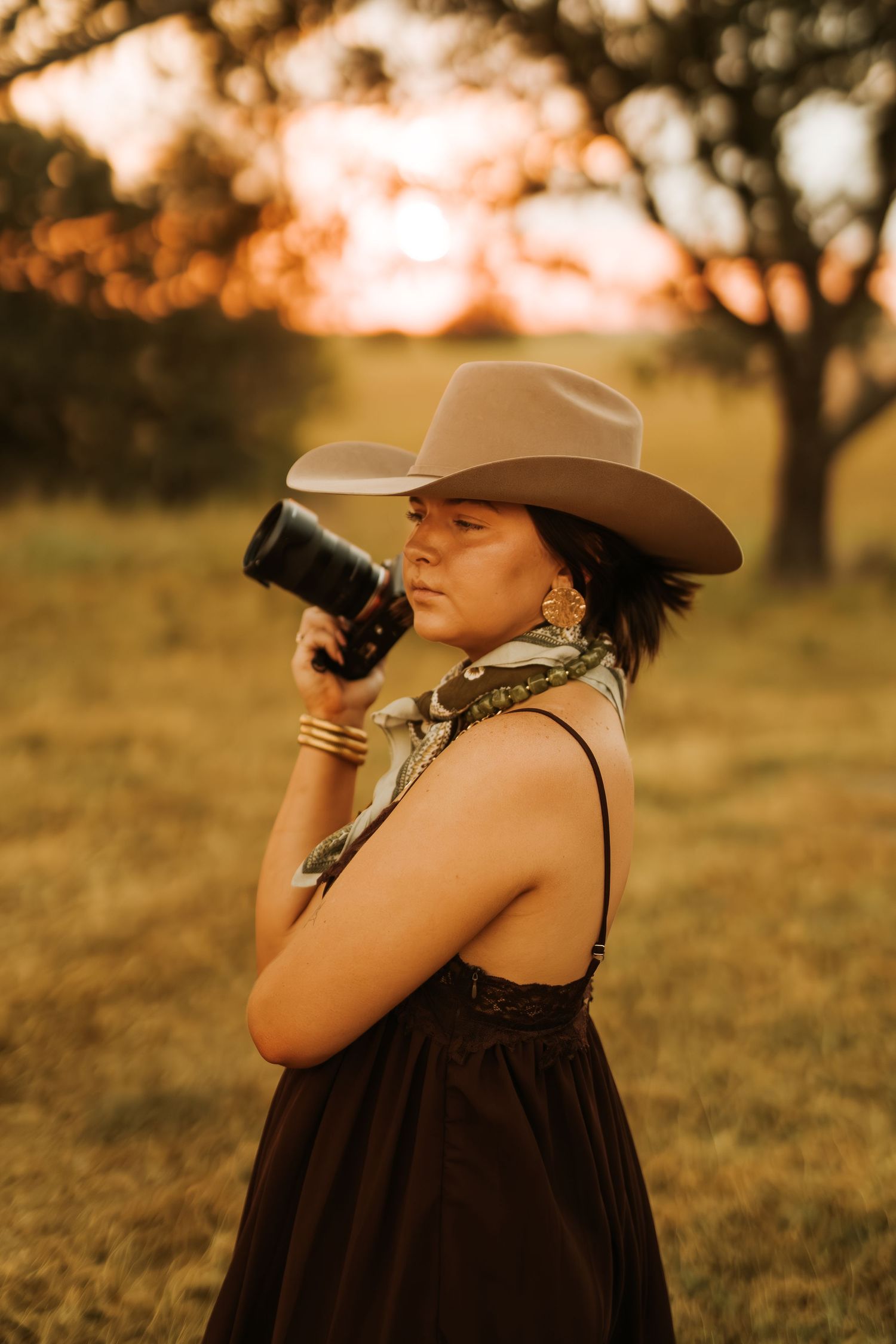 Woman in cowboy hat and black dress photographing with camera lens at golden hour sunset in a field.