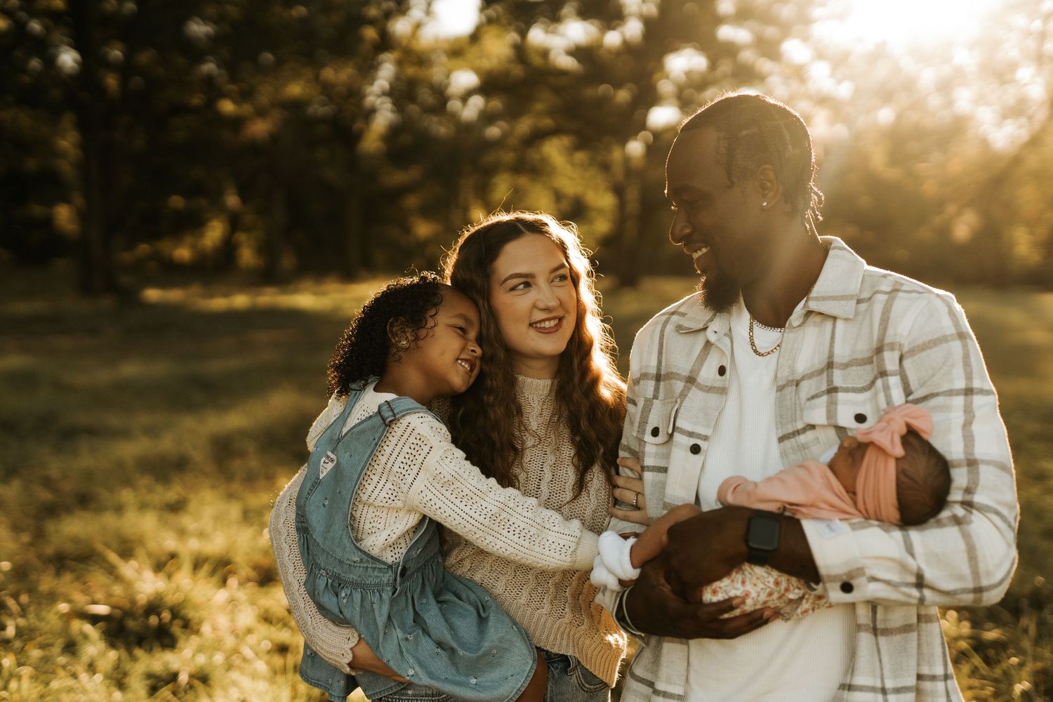 A happy interracial family with a young girl and newborn baby enjoying a golden hour outdoor photo session.