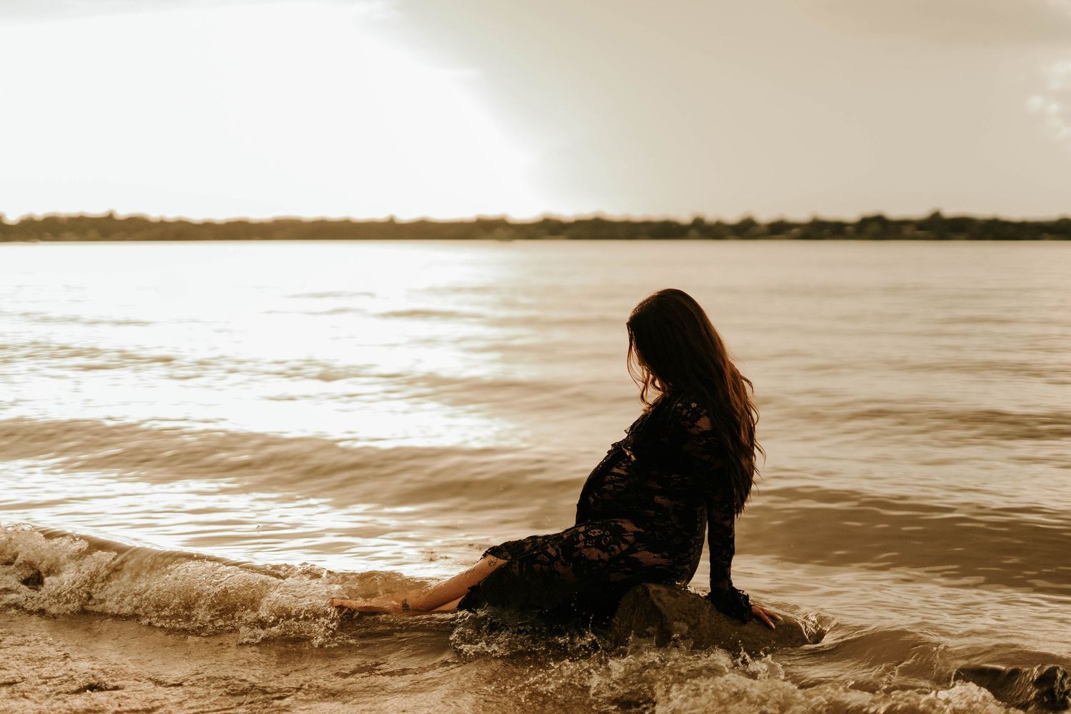 Woman in black dress sitting on sandy beach shore with waves washing over her feet at golden sunset.
